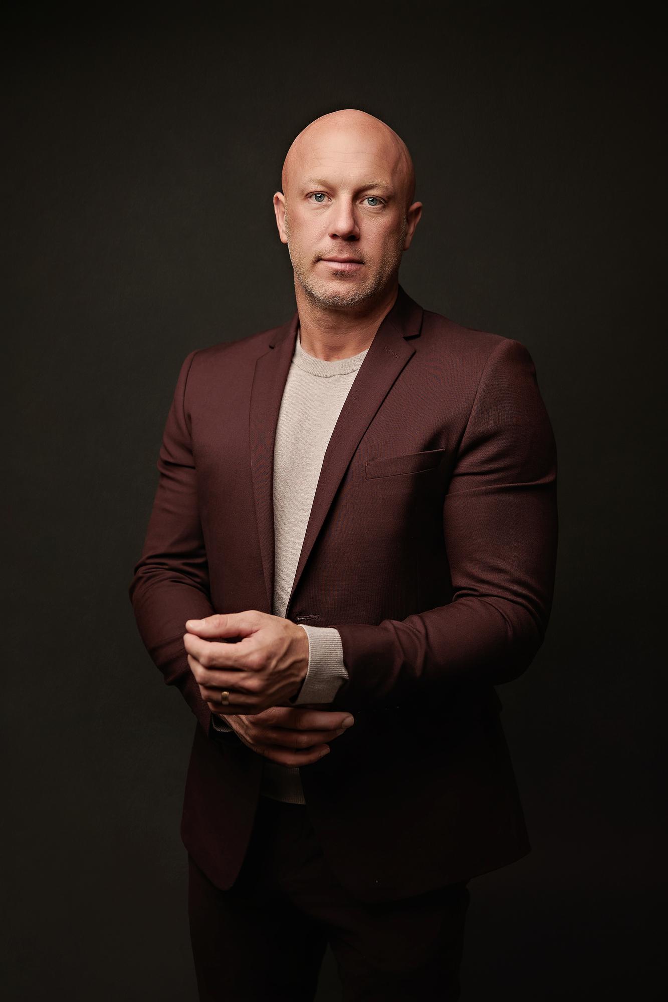 Professional headshot Calgary studio: Authoritative male executive in a deep burgundy blazer and grey t-shirt standing with hands clasped in front, dark charcoal studio background