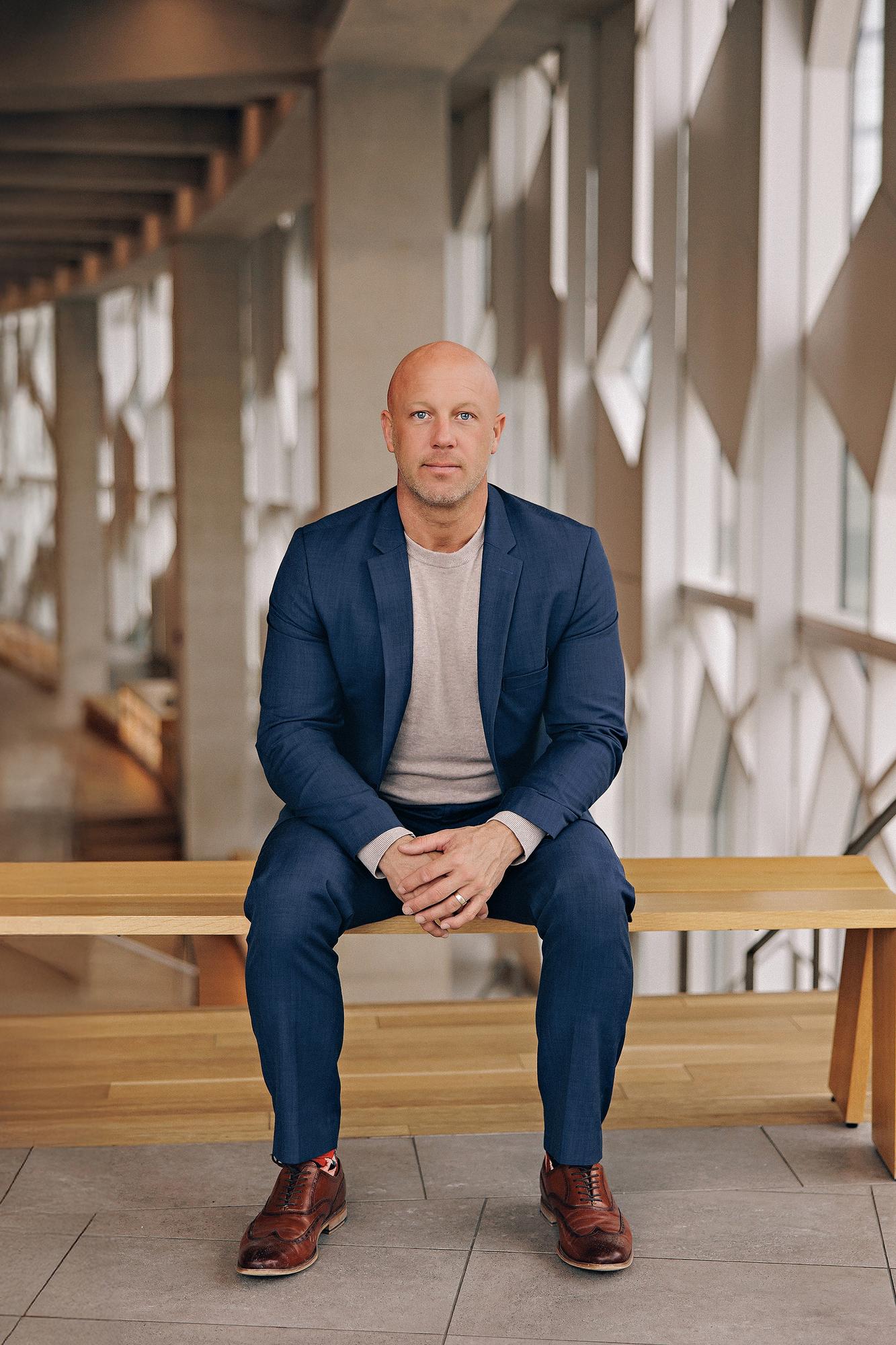 Environmental personal branding headshot Calgary: Authoritative male executive in a navy blue blazer and grey t-shirt seated on a wood bench with hands clasped inside a contemporary architectural atrium with geometric windows