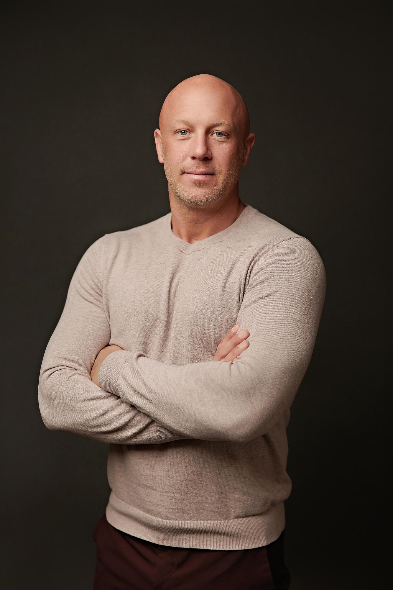 Professional headshot Calgary studio: Approachable male professional in a sand beige crewneck sweater standing with arms crossed and a relaxed smile against a dark charcoal background