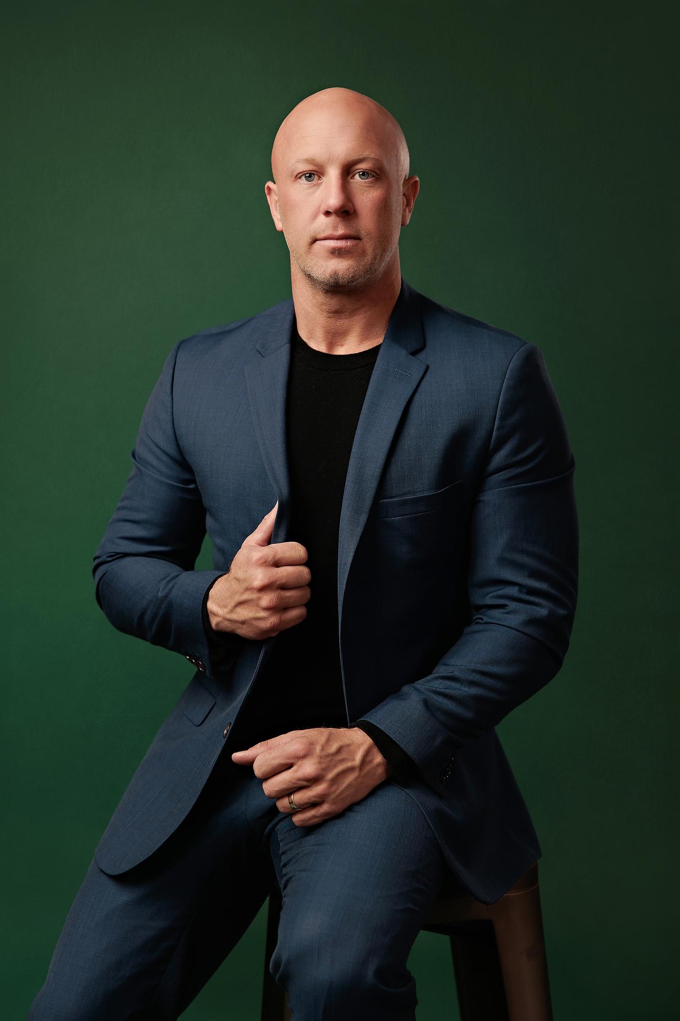 Professional headshots Calgary studio: Powerful male executive in a navy blue blazer and black t-shirt seated on a stool with one hand gripping his lapel, deep emerald green studio background