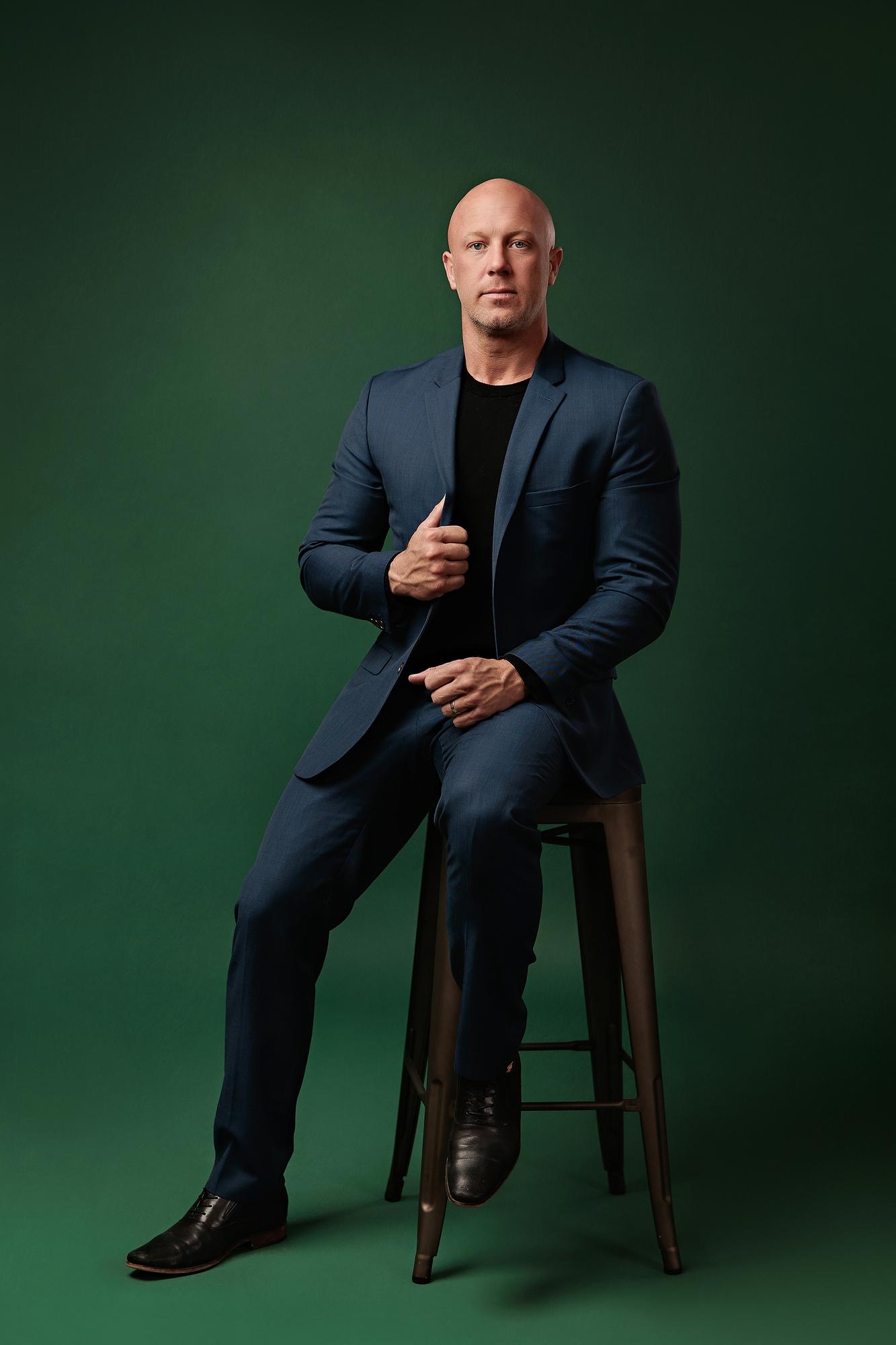 Full-length professional headshot Calgary studio: Confident male professional in a navy blue suit and black t-shirt seated on a metal barstool with hand on lapel and black leather dress shoes, emerald green background