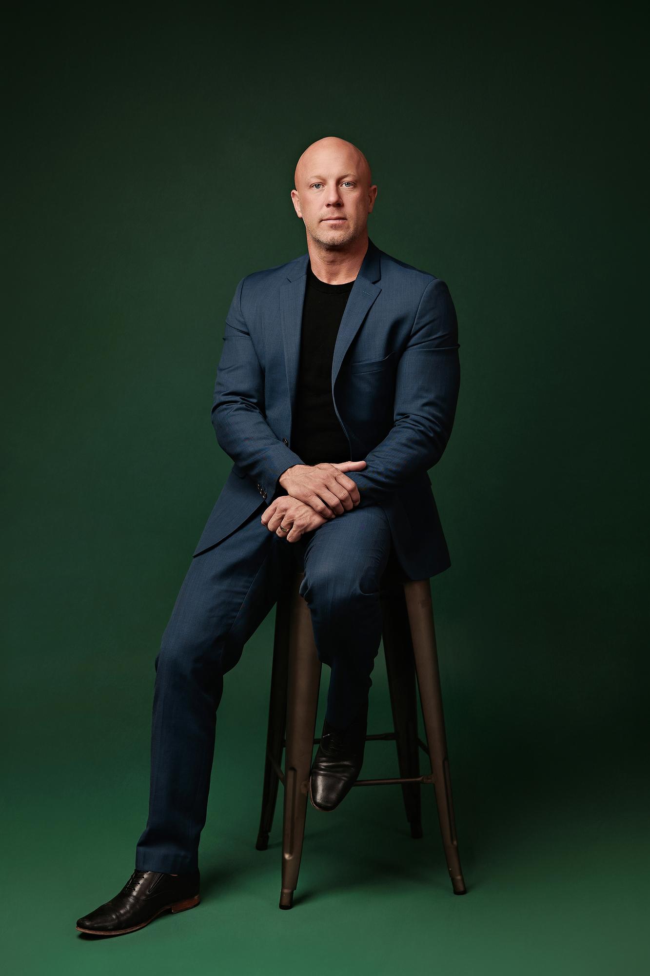 Full-length professional headshot Calgary studio: Commanding male professional in a navy blue suit and black t-shirt seated on a metal barstool with hands clasped and black dress shoes, emerald green background