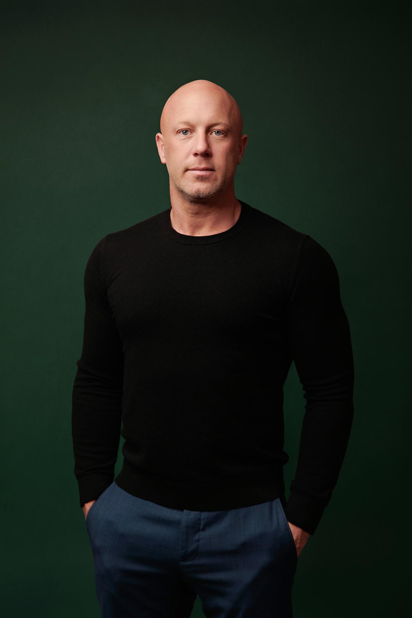 Professional headshot Calgary studio: Intense male professional in a fitted black crewneck sweater and navy trousers standing with hands in pockets against a deep emerald green background