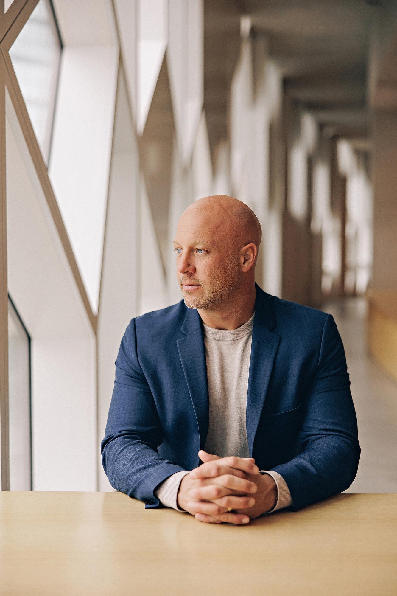 On-location personal branding photography Calgary: Thoughtful male professional in a navy blue blazer and grey t-shirt seated at a wood table with hands clasped, gazing away down a light-filled architectural corridor