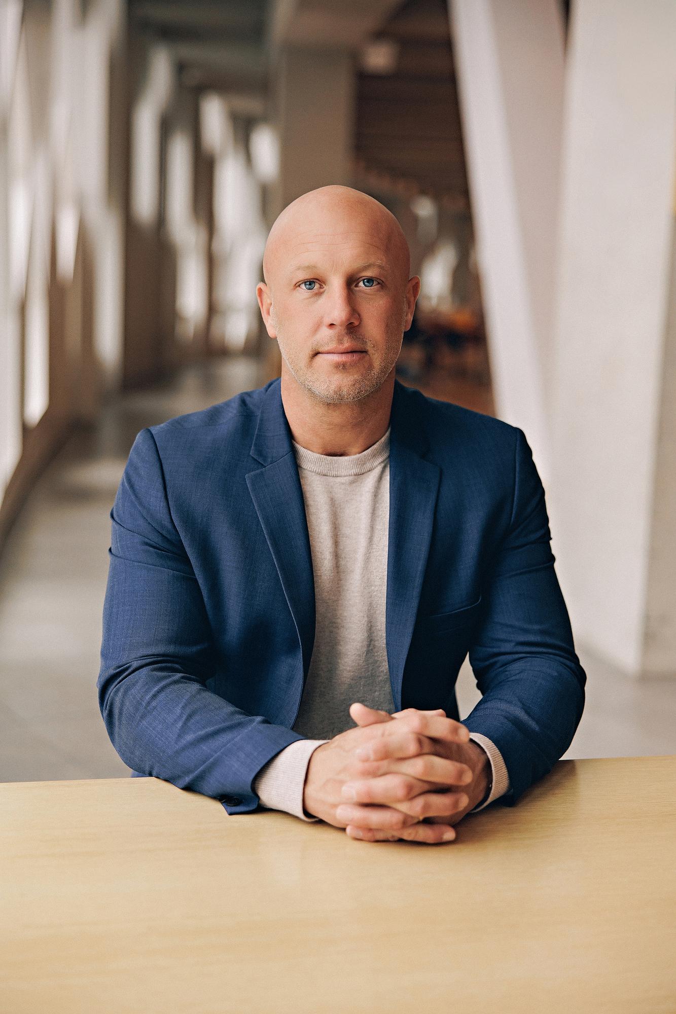 Personal branding headshot Calgary: Focused male executive in a navy blue blazer and grey t-shirt seated at a wood table with hands clasped, looking directly at camera in a bright modern corridor