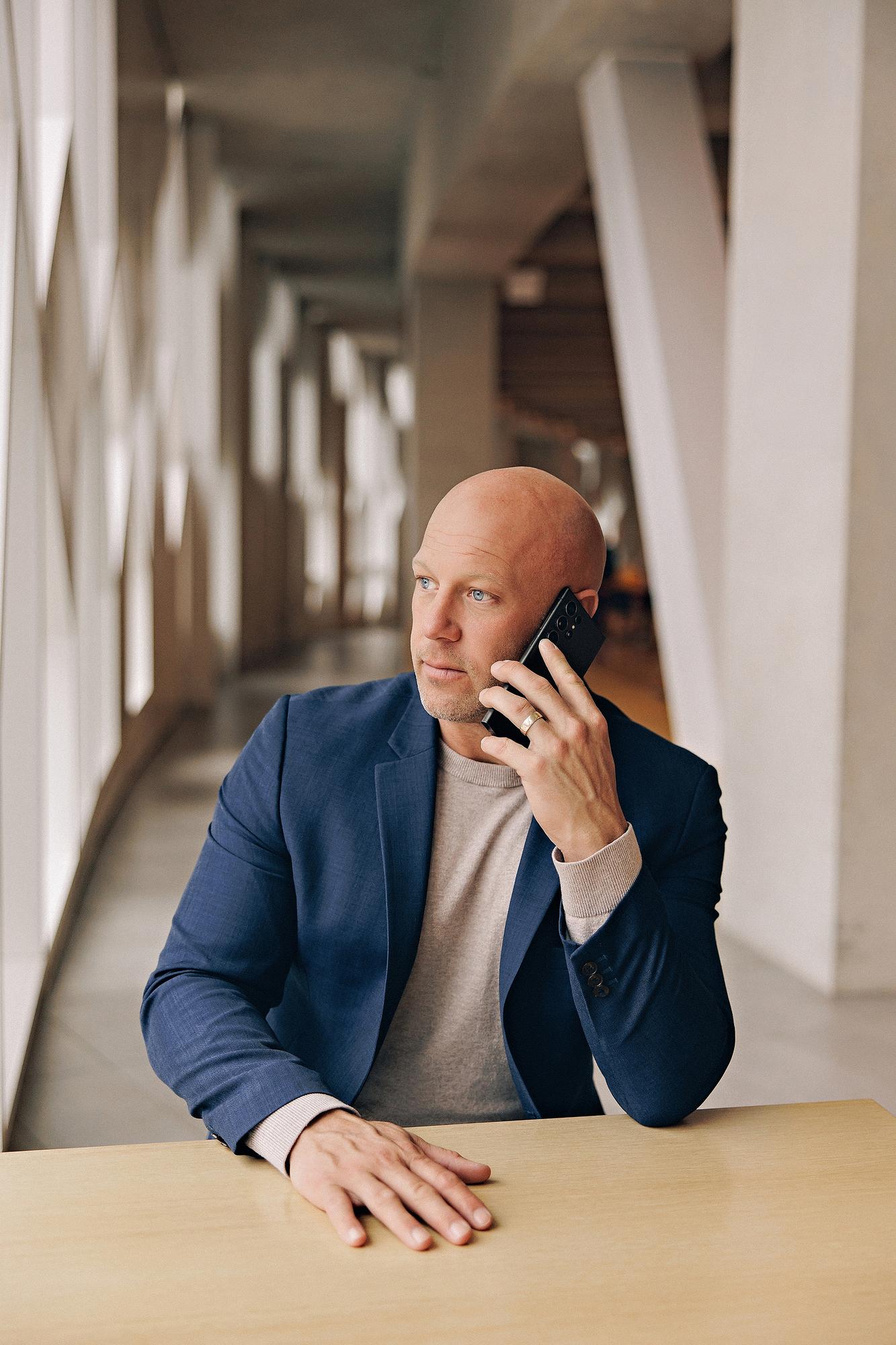 Lifestyle personal branding photography Calgary: Professional man in a navy blue blazer and grey t-shirt seated at a wood table talking on a smartphone, gazing away in a sunlit architectural corridor