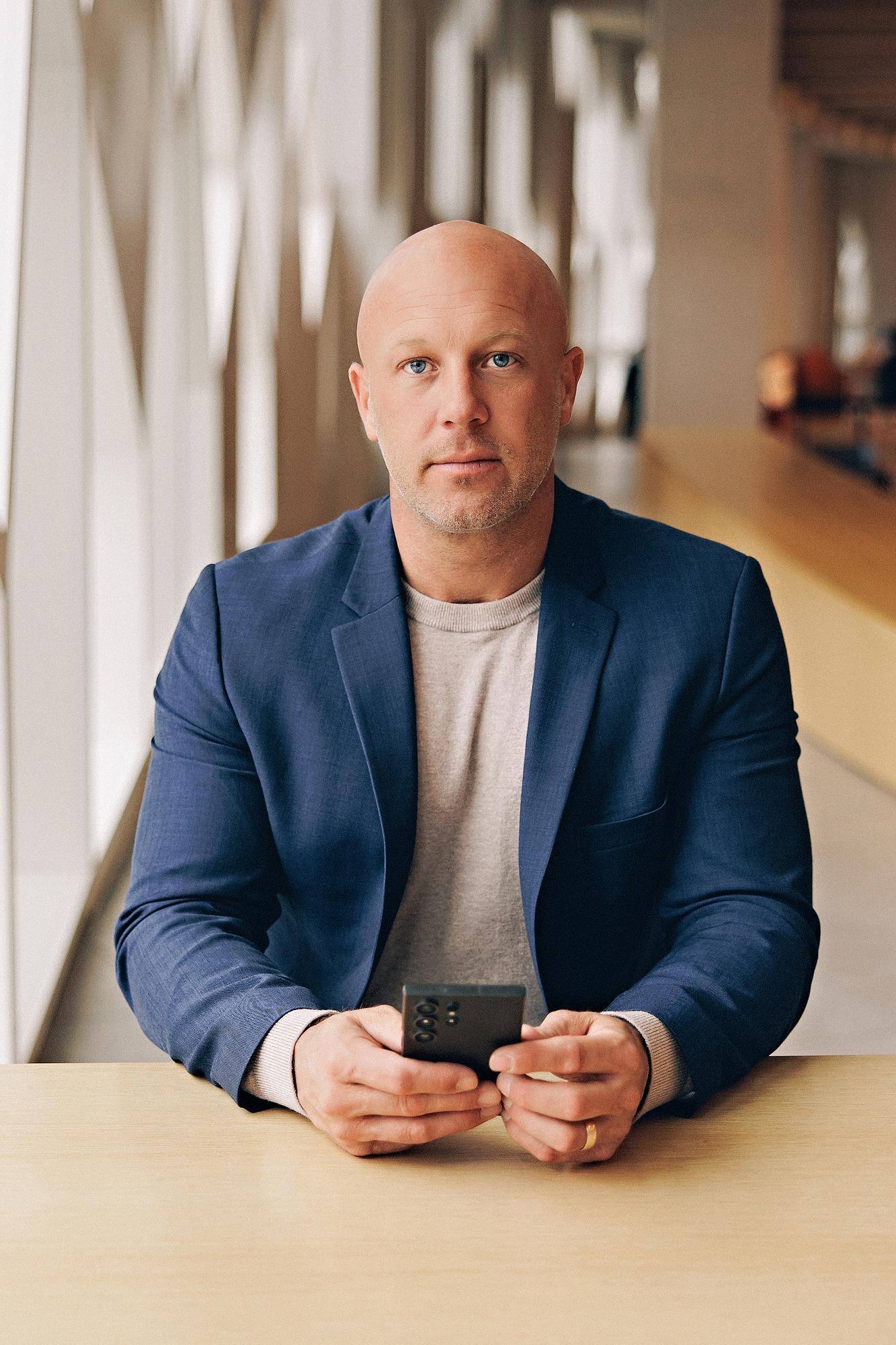 Personal branding session Calgary: Intense male professional in a navy blue blazer and grey t-shirt seated at a wood table holding a smartphone and looking directly at camera in a bright modern office corridor