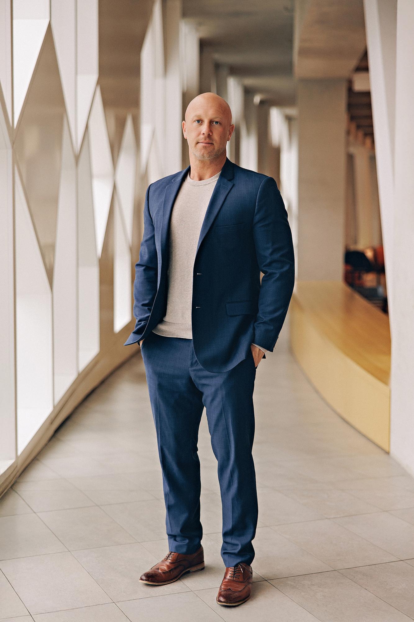 Full-length personal branding portrait Calgary: Confident professional man in a navy blue suit and grey t-shirt with hands in pockets standing in a bright architectural corridor with geometric windows and brown leather oxfords