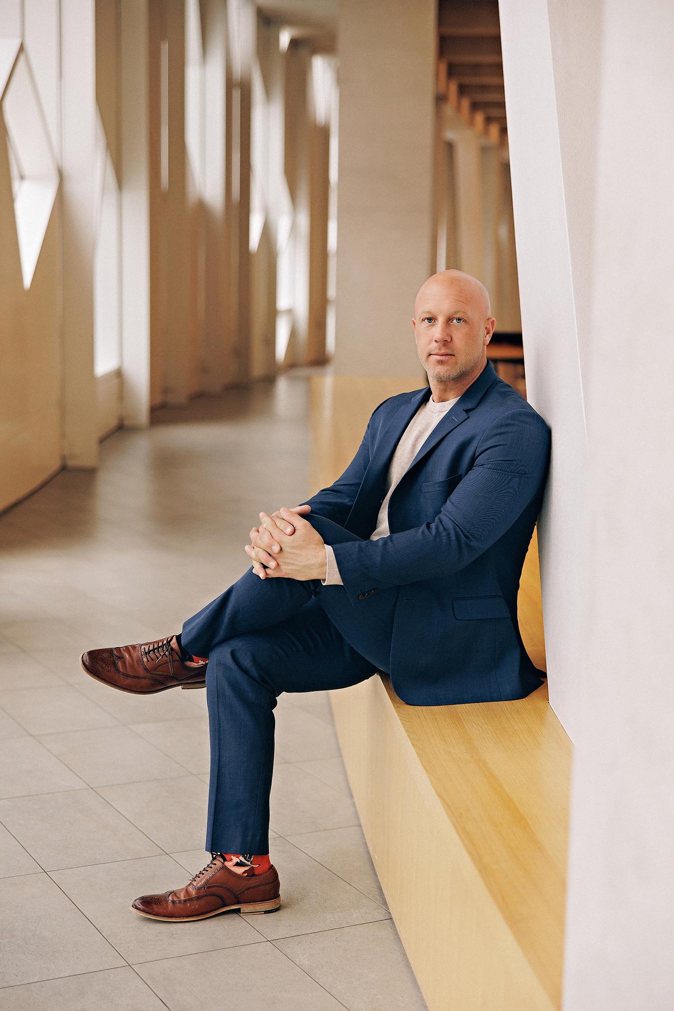 On-location personal branding photography Calgary: Relaxed male executive in a navy blue suit and grey t-shirt seated on a wood ledge with one leg crossed and hands clasped in a modern architectural corridor with geometric windows