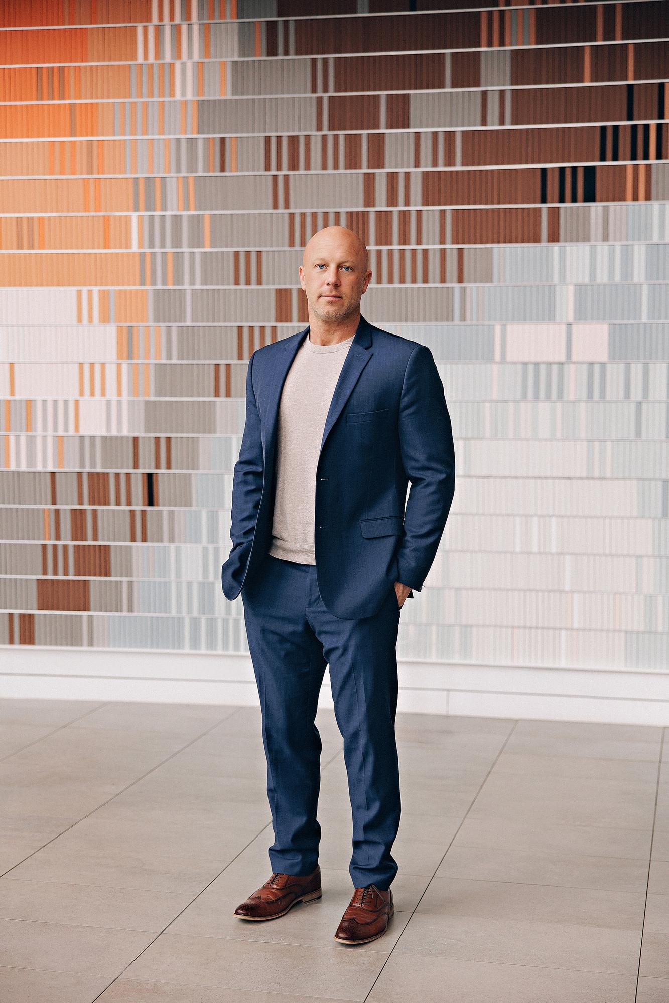 Full-length personal branding portrait Calgary: Confident professional man in a navy blue suit and grey t-shirt standing with hands in pockets in front of a large colourful striped art installation inside a Calgary office building