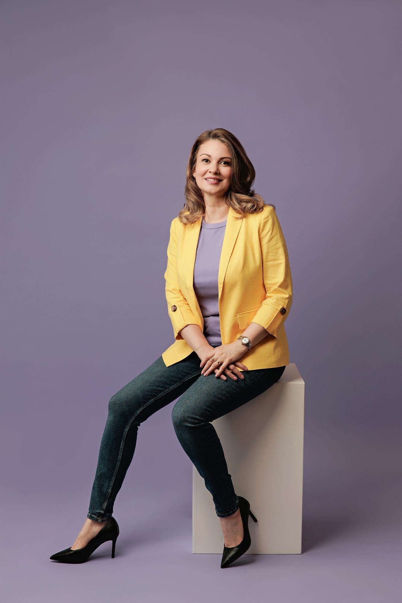 Personal branding photography Calgary studio: Woman in a black blazer and mustard yellow turtleneck seated on a stool with a composed expression, warm golden amber background