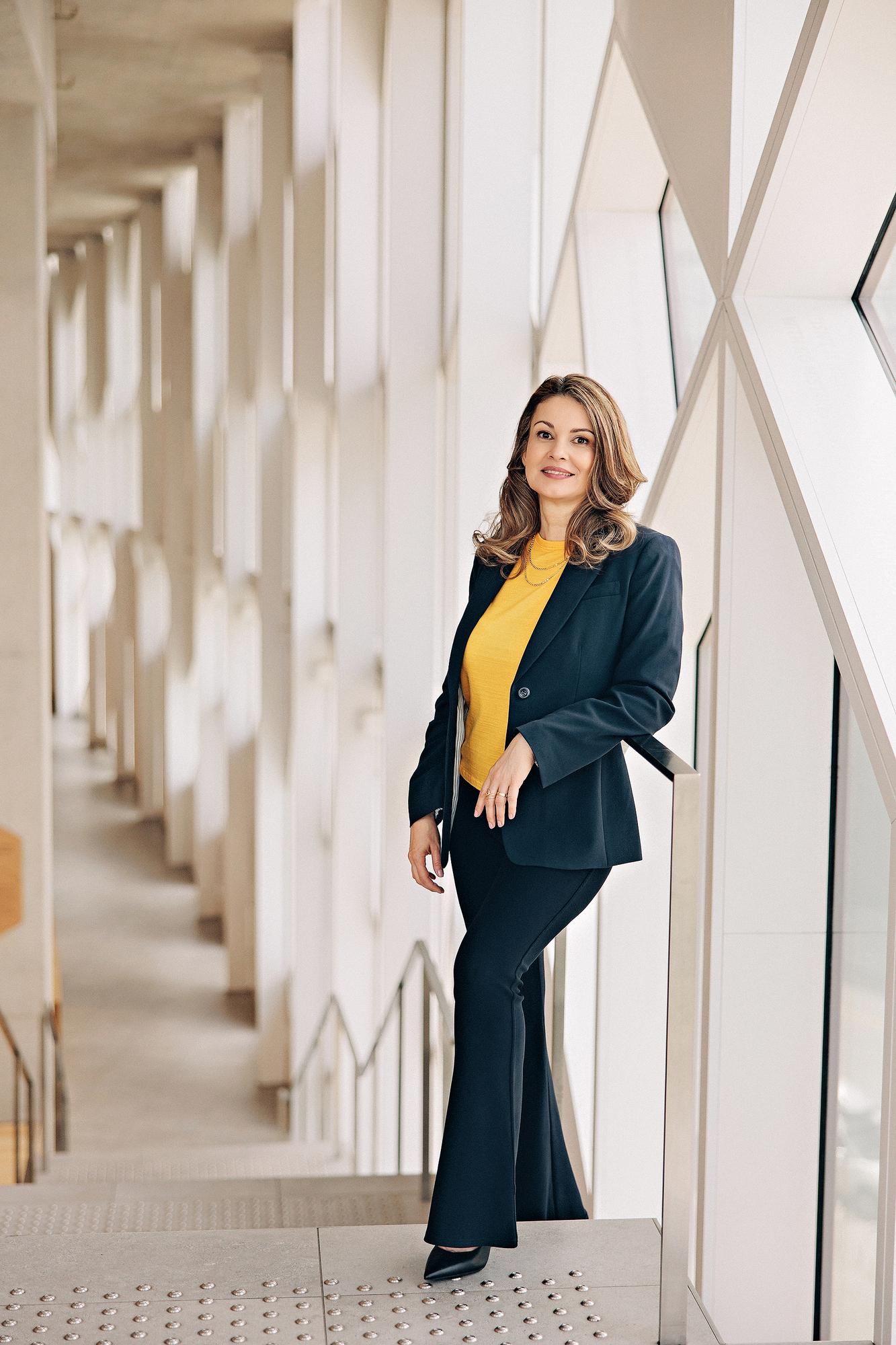 Editorial personal branding photography Calgary: Dramatic full-length portrait of a woman in a black leather moto jacket and black turtleneck seated on a stool gazing away, dark background