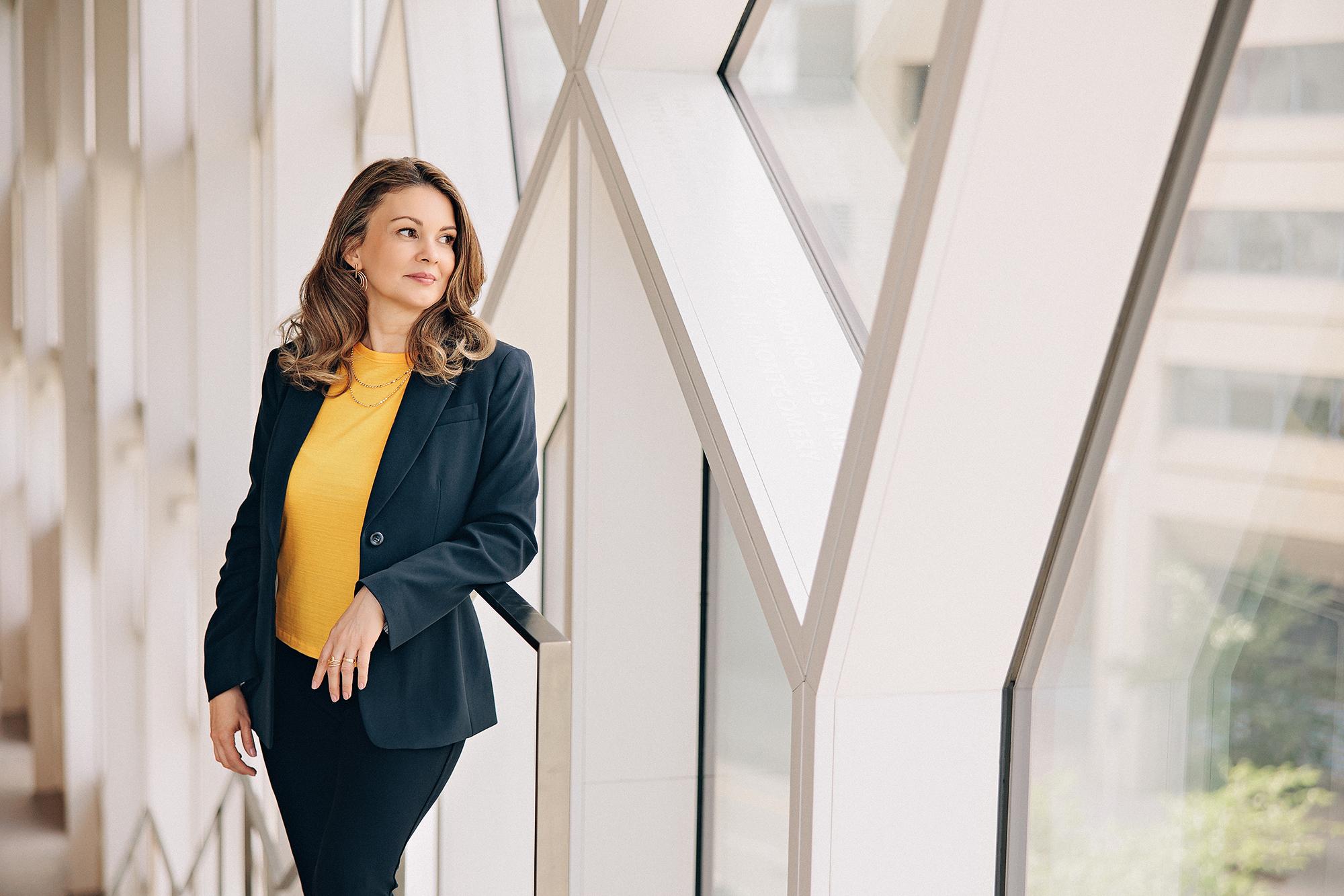 On-location personal branding headshot Calgary: Confident woman in a navy blazer and mustard yellow top leaning on a railing in a bright modern architectural corridor