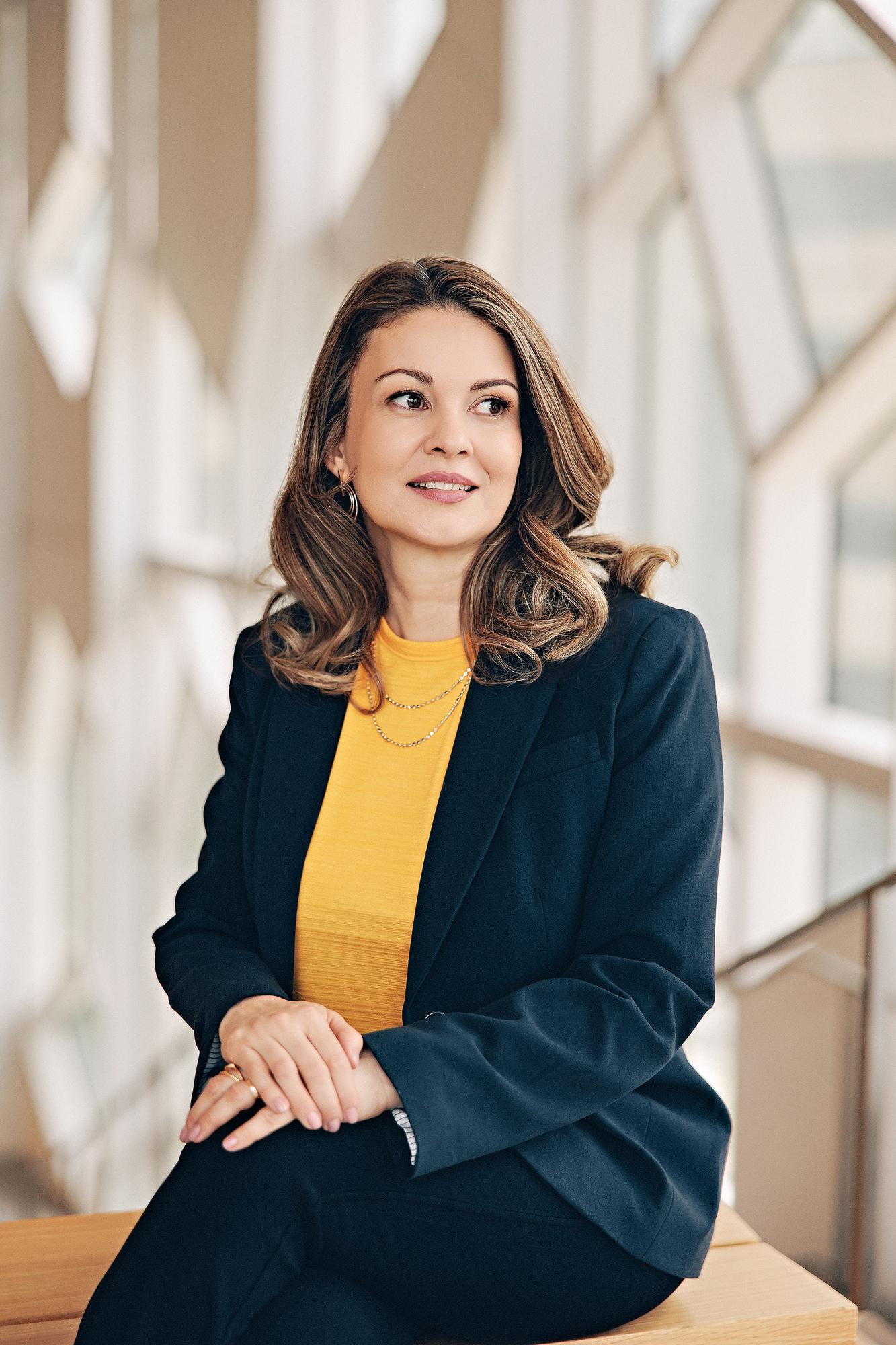 Environmental personal branding photography Calgary: Thoughtful woman in a navy blazer and mustard yellow top leaning on a railing gazing sideways beside geometric windows in a modern Calgary building