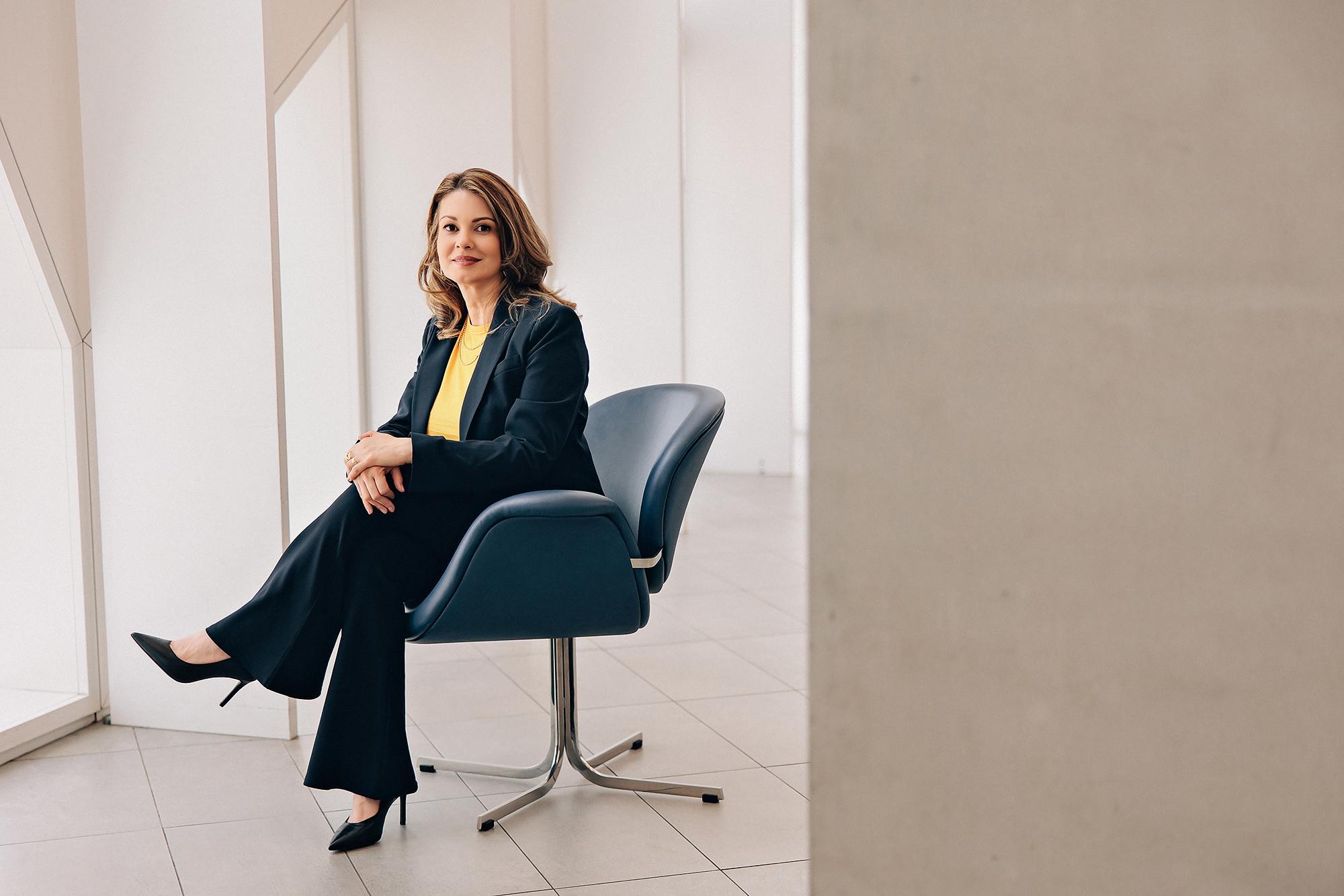 On-location personal branding photography Calgary: Businesswoman in a navy suit and golden yellow top seated on a wood bench with hands clasped, gazing upward with a serene expression in a bright architectural atrium