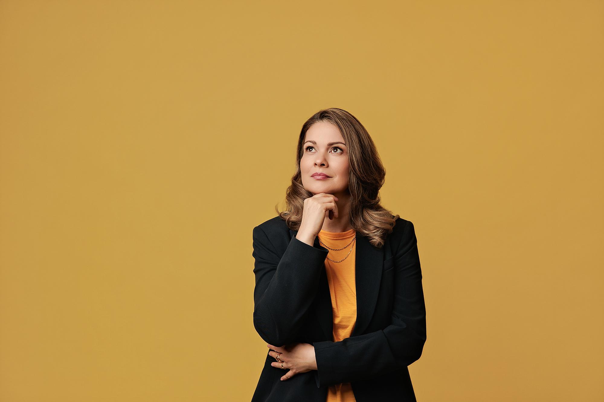 Environmental personal branding photography Calgary: Composed woman in a navy blazer and mustard yellow top seated in a sculptural blue lounge chair in a bright modern interior