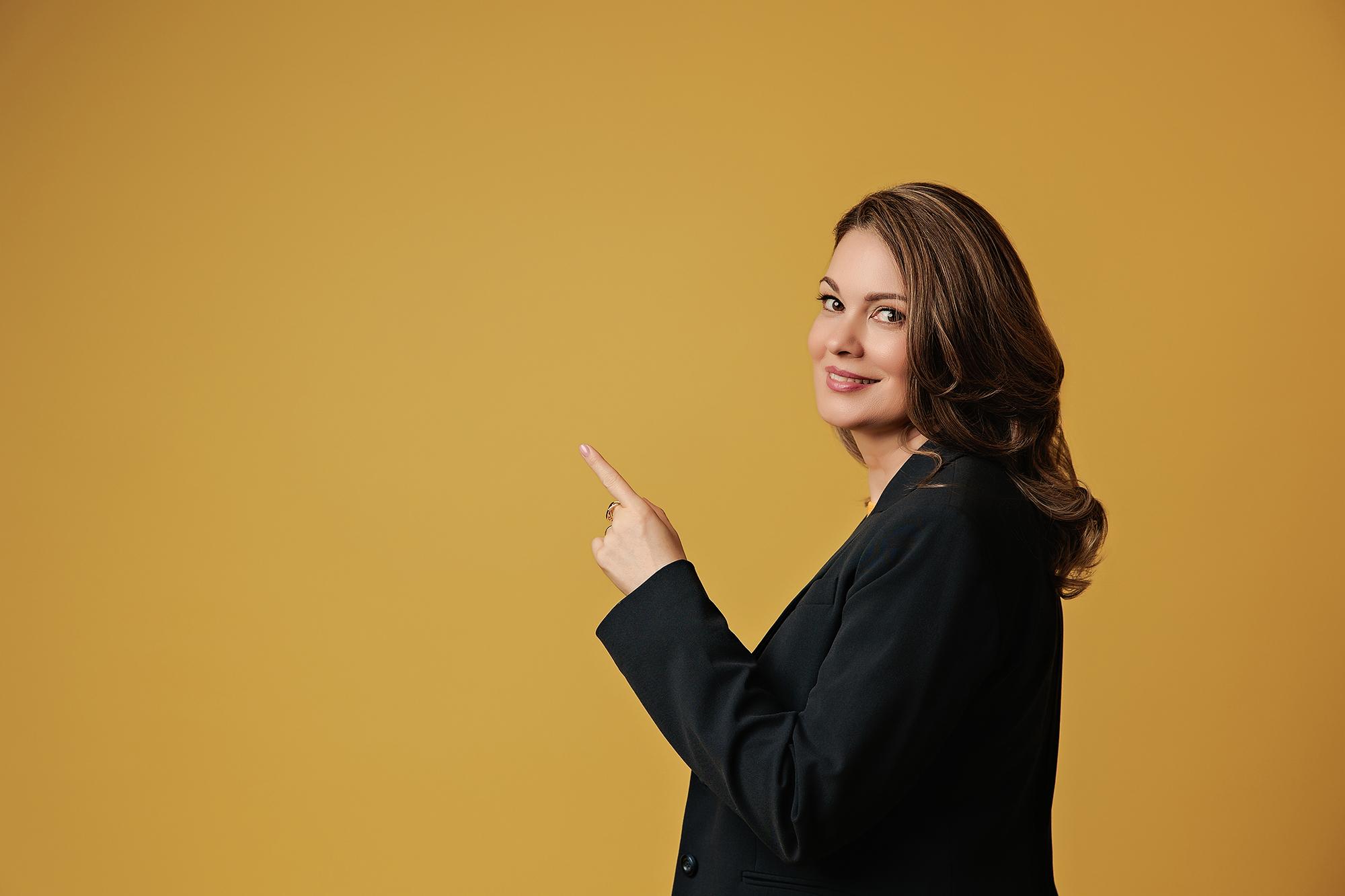 Personal branding headshot Calgary studio: Playful female entrepreneur in a black blazer turning over her shoulder and pointing to the side with a coy smile against a warm golden amber background
