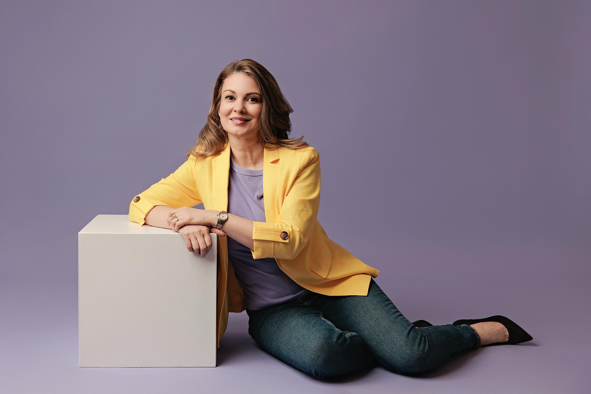Personal branding photography Calgary studio: Relaxed woman in a yellow blazer and lavender top seated on the floor leaning against a white cube, soft lavender background