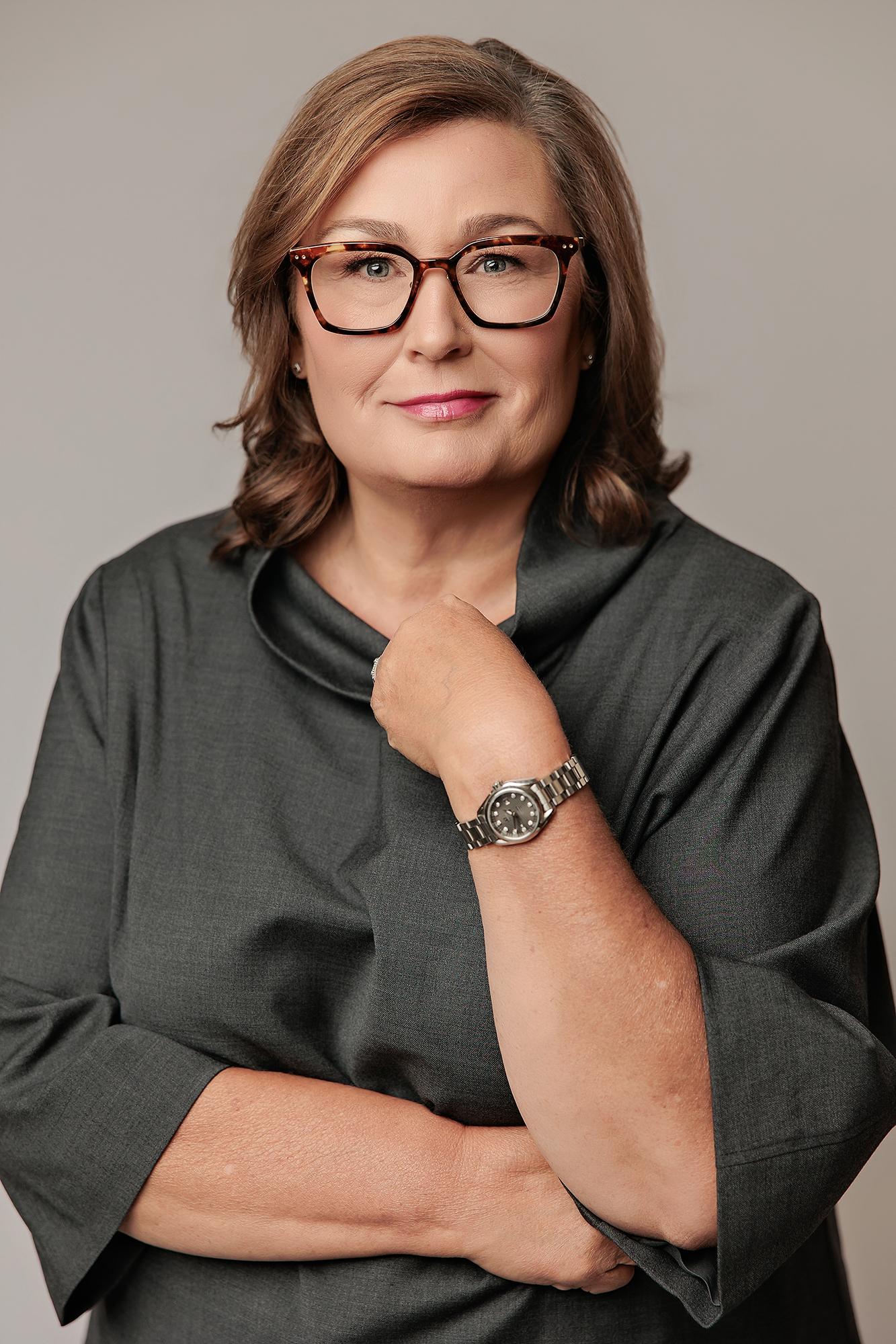 A professional woman wearing glasses and a charcoal top poses for a Calgary headshot photographer, showcasing clean studio lighting and a calm expression.