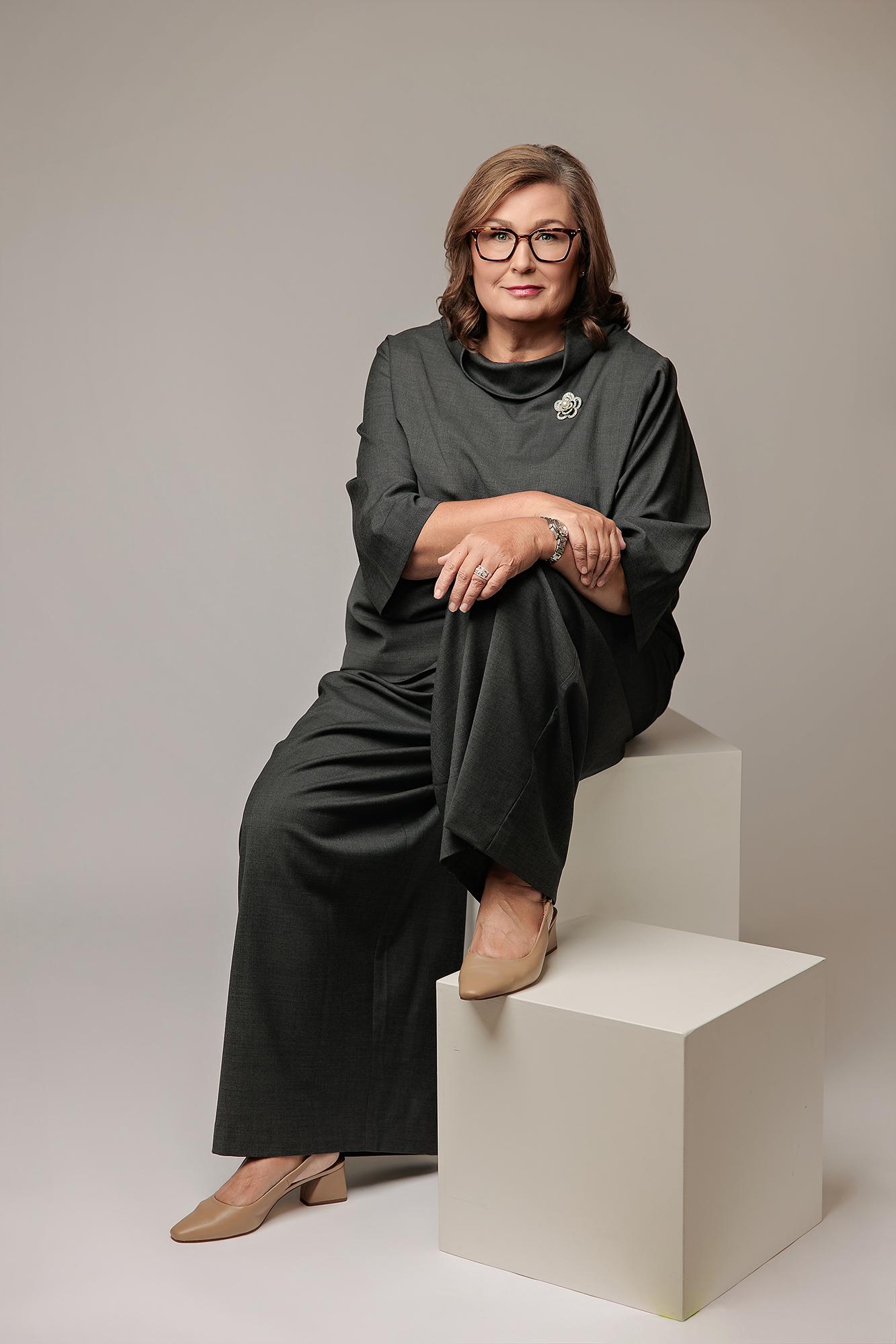 A professional woman in a dark grey suit sits confidently on a white cube for this Calgary studio headshot, showcasing elegant personal branding.