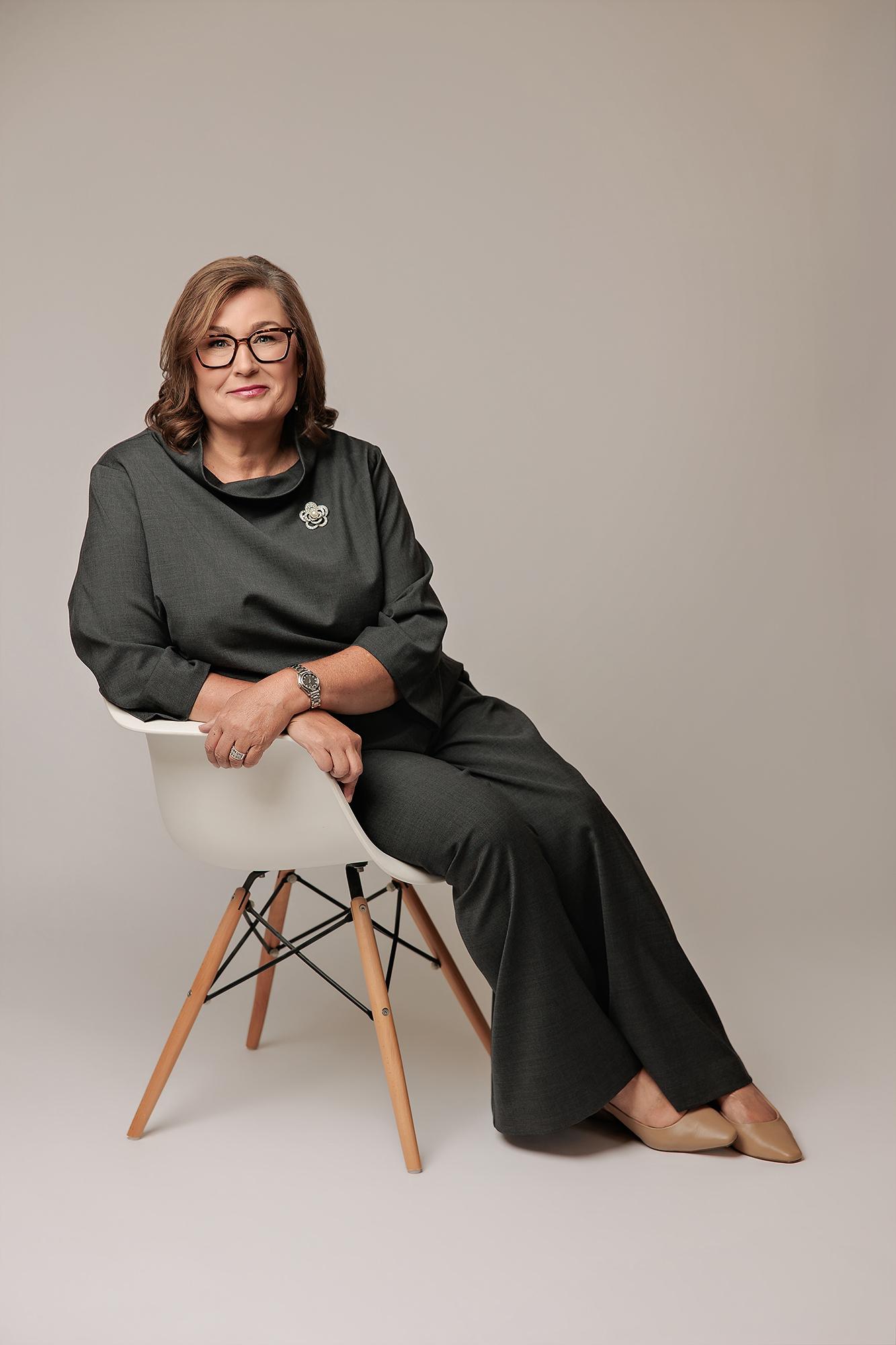 A professional woman in a charcoal grey outfit sits confidently on a white chair, showcasing the polished style of our Calgary headshot studio.