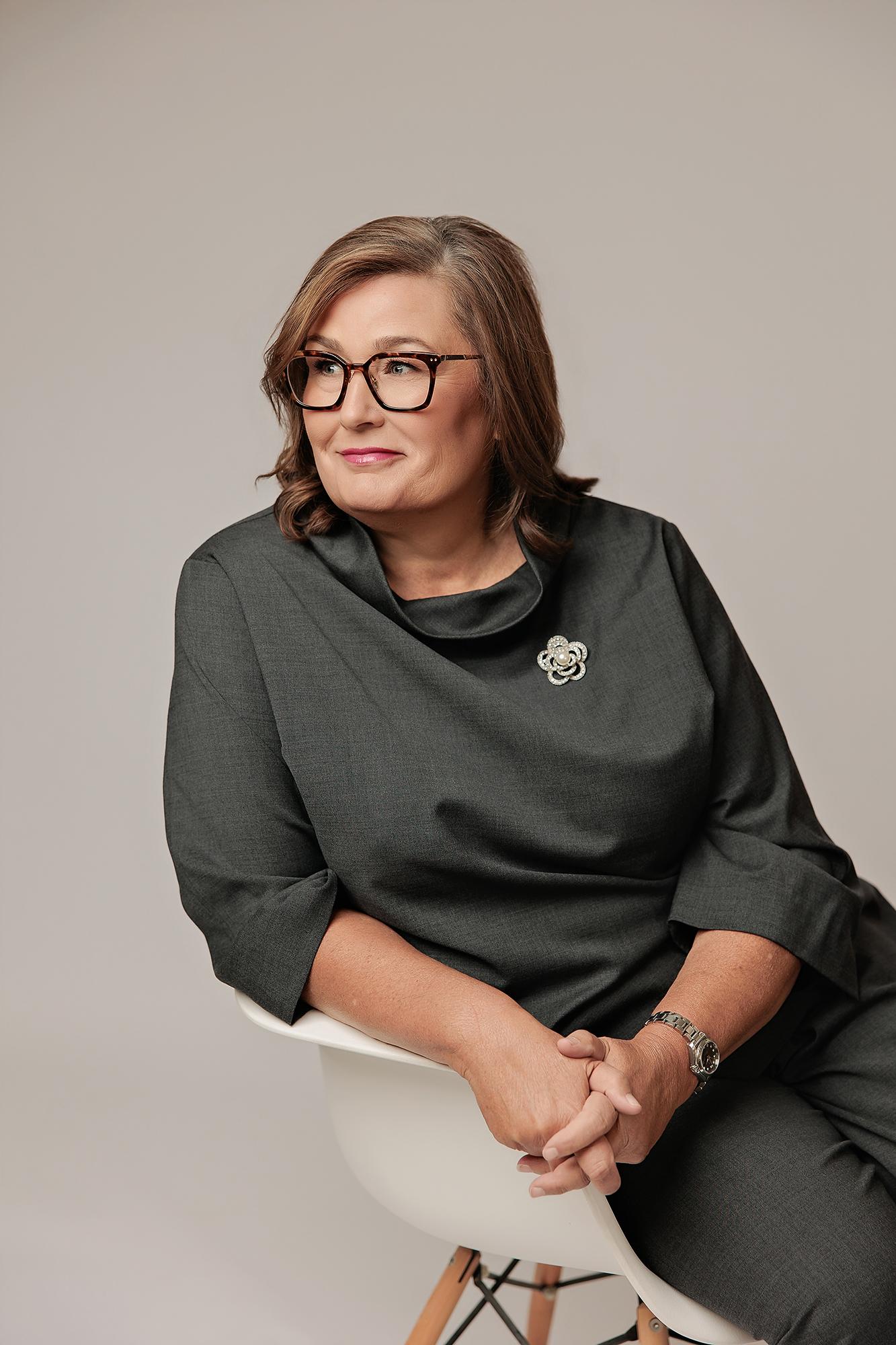 A professional woman with glasses and a charcoal dress poses for a Calgary headshot session, showcasing soft studio lighting and a confident expression.