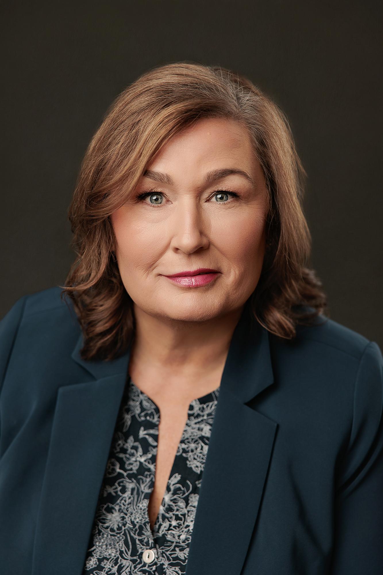 A middle-aged woman with a warm expression wears a teal blazer in this professional headshot captured at a Calgary photography studio.