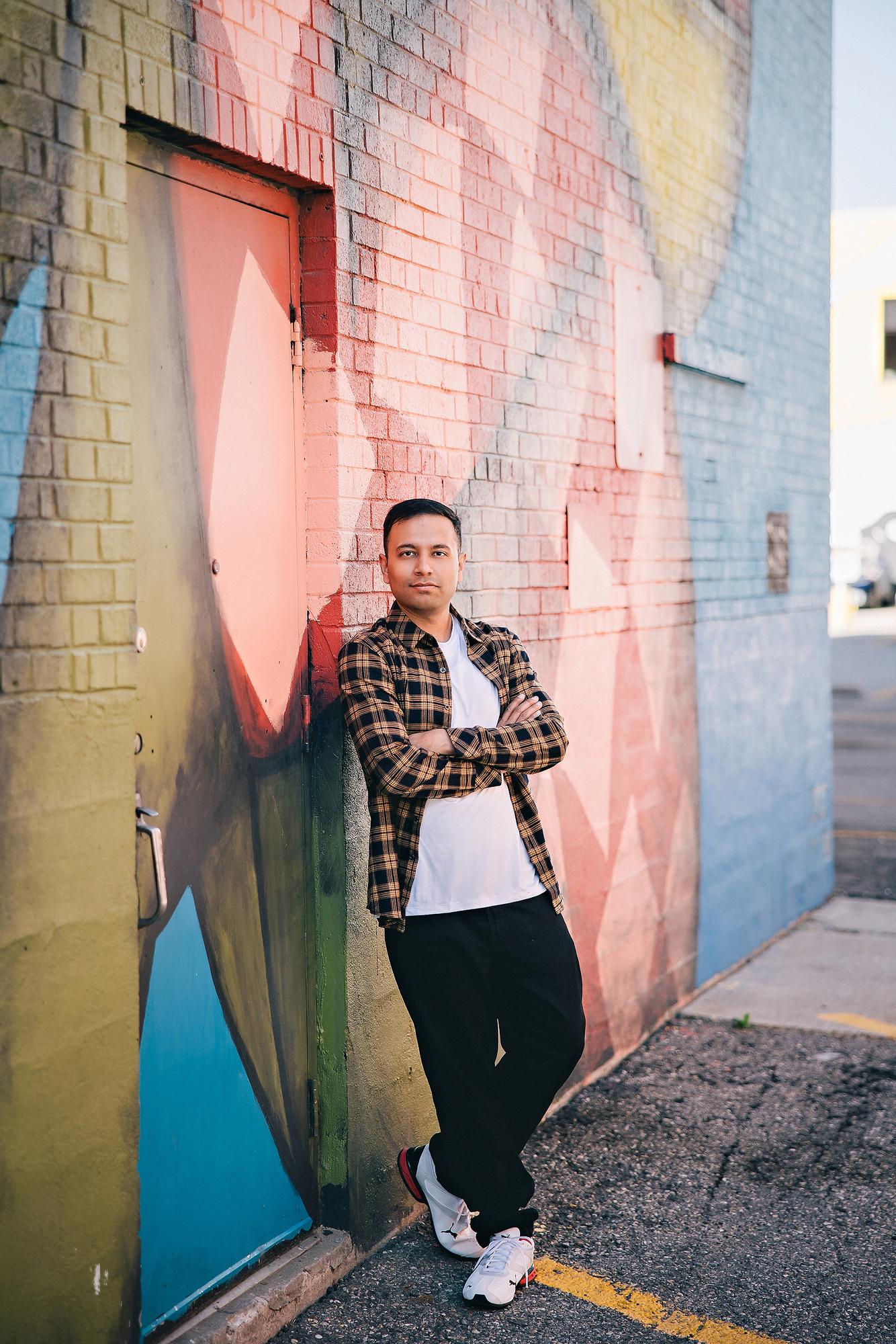 Urban personal branding headshot Calgary: Confident young man in a tan plaid flannel shirt and white tee standing with arms crossed against a vibrant colourful street mural