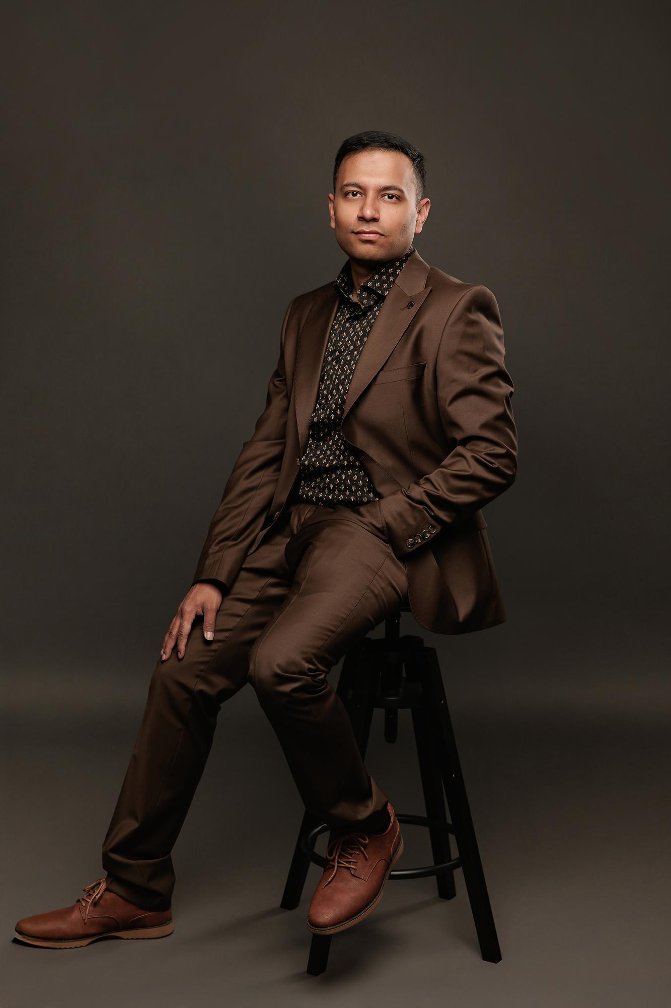 Executive headshot Calgary studio: Intense man in a rich brown suit and black gold-patterned shirt seated on a black stool with one leg raised and a direct gaze, dark background