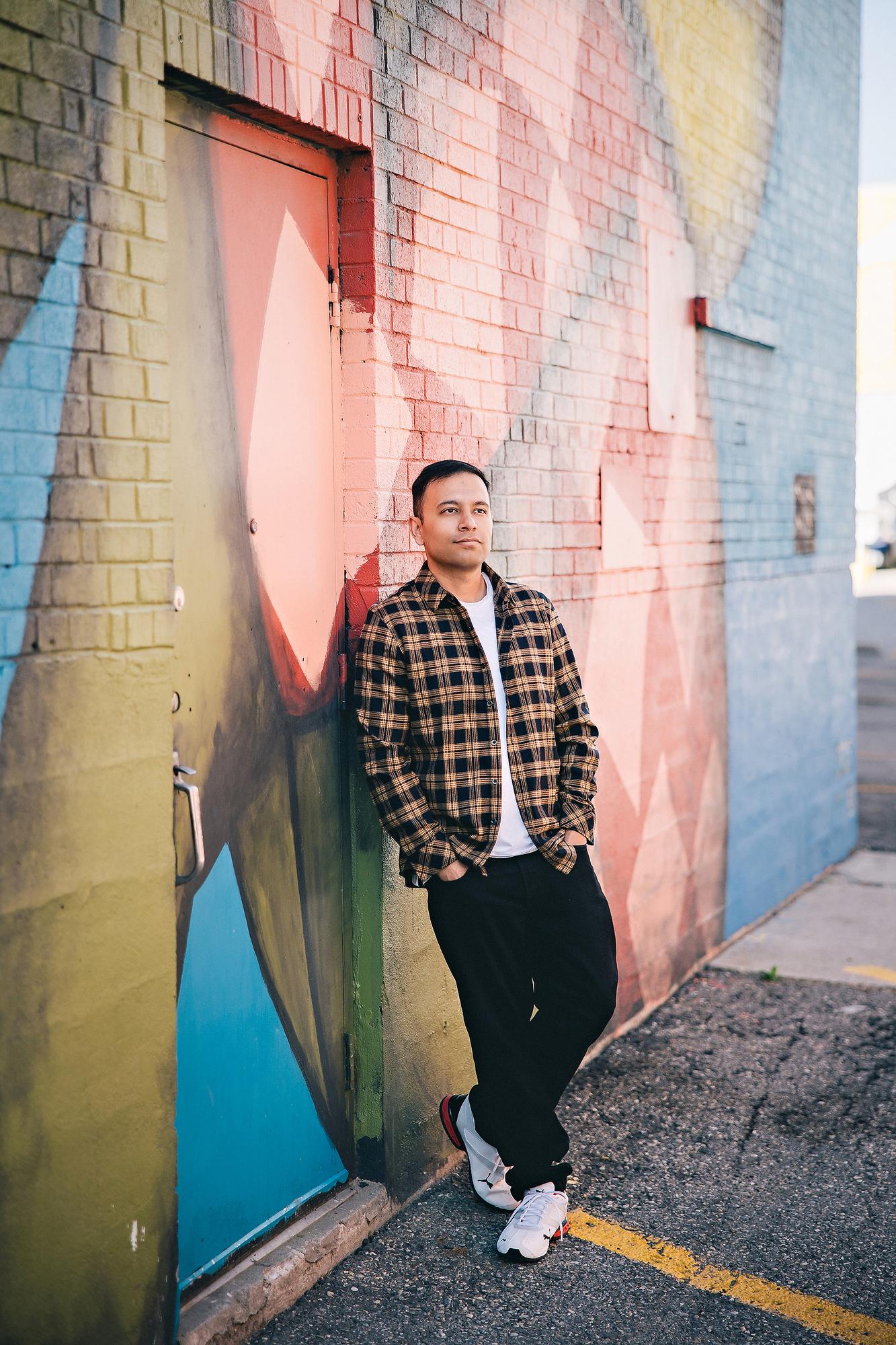 Outdoor personal branding photography Calgary: Relaxed young man in a tan plaid flannel shirt and white tee leaning with hands in pockets against a bright colourful street mural