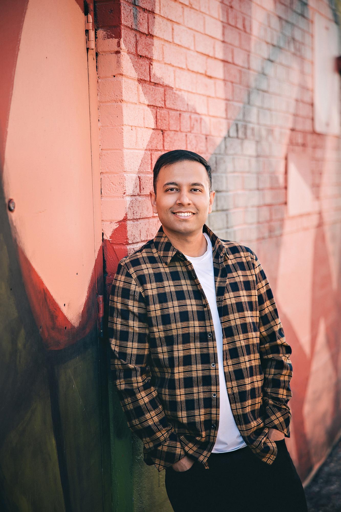 Urban personal branding headshot Calgary: Smiling young man in a tan plaid flannel shirt and white tee with hands in pockets leaning against a colourful street mural