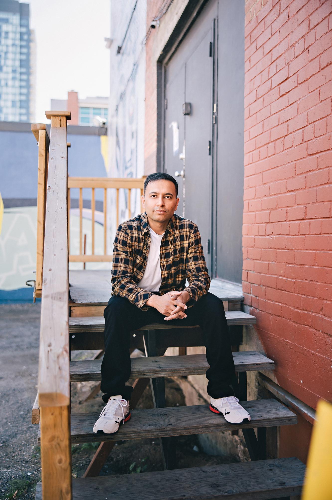 On-location personal branding photography Calgary: Relaxed young man in a tan plaid flannel shirt seated on wooden back-alley stairs with hands clasped, red brick wall and Calgary skyline behind him