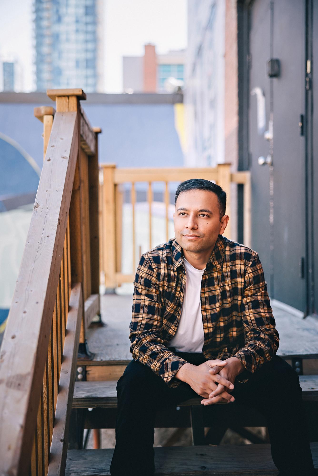 Outdoor personal branding headshot Calgary: Thoughtful young man in a tan plaid flannel shirt seated on wooden stairs gazing sideways with the Calgary downtown skyline in the background