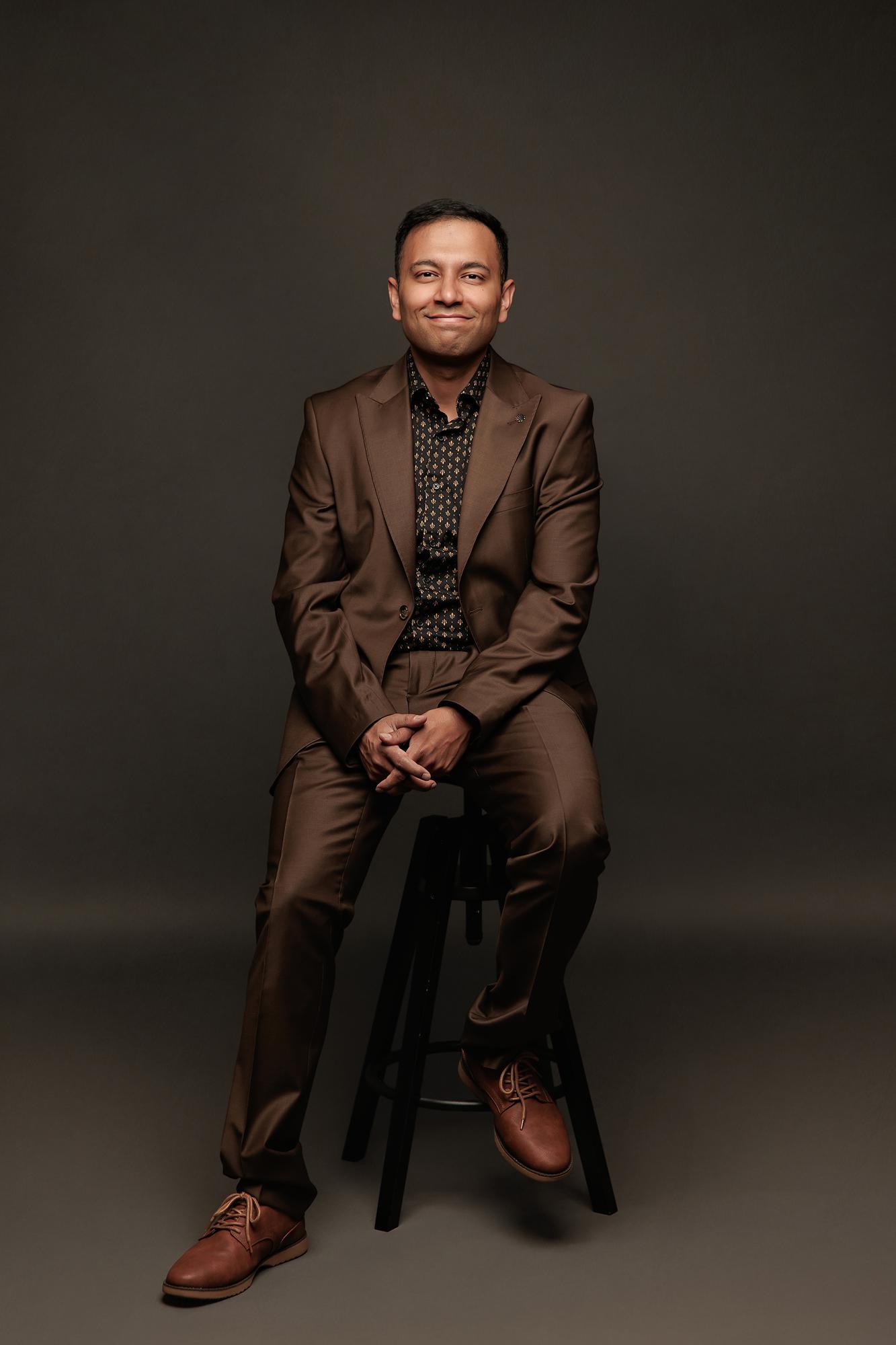 Professional headshot Calgary studio: Smiling man in a rich brown suit and black gold-patterned shirt seated on a black stool with hands clasped against a dark background