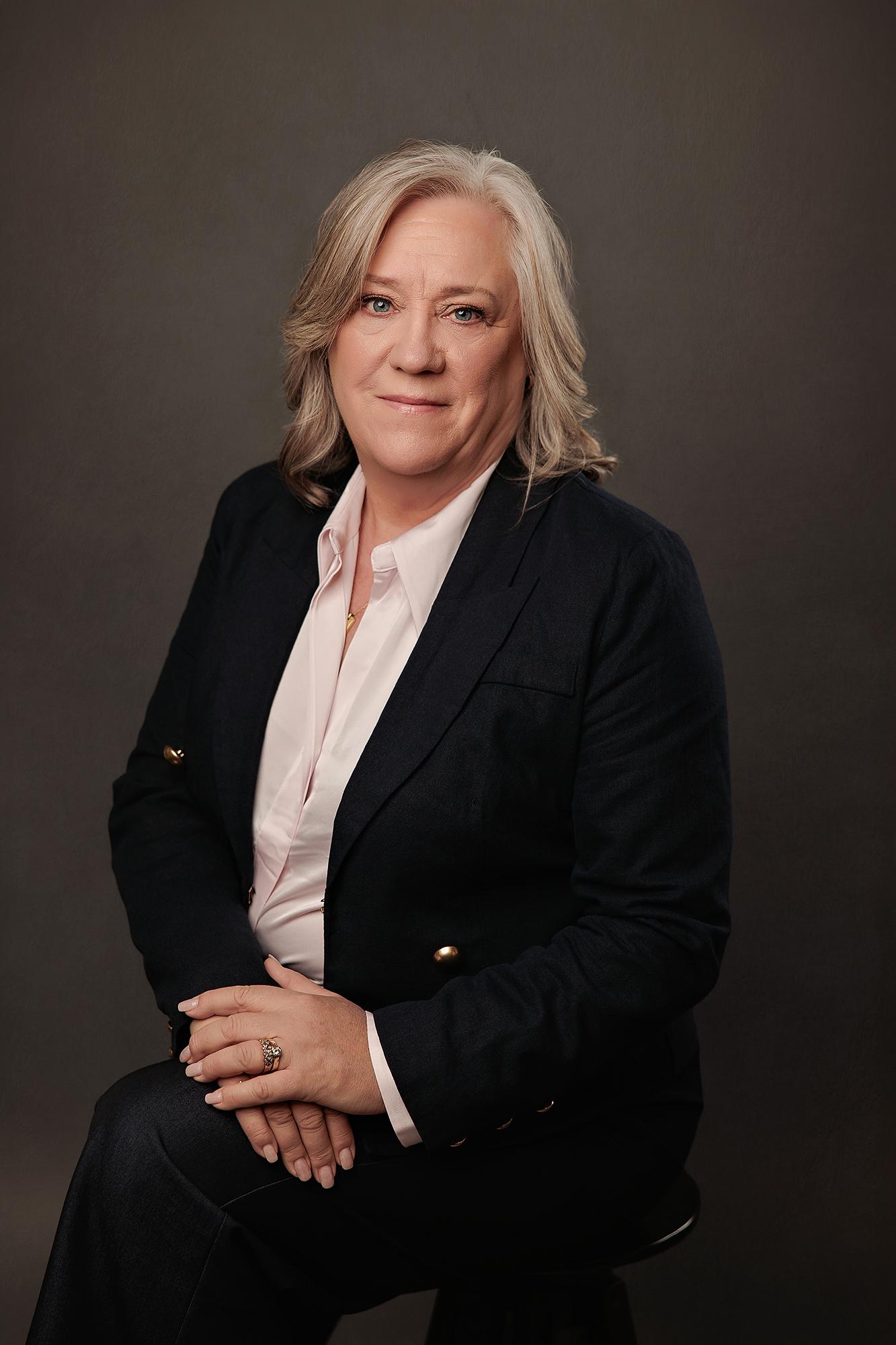 A professional woman with blonde hair wears a navy blazer in this Calgary studio headshot, captured with soft lighting against a dark backdrop.