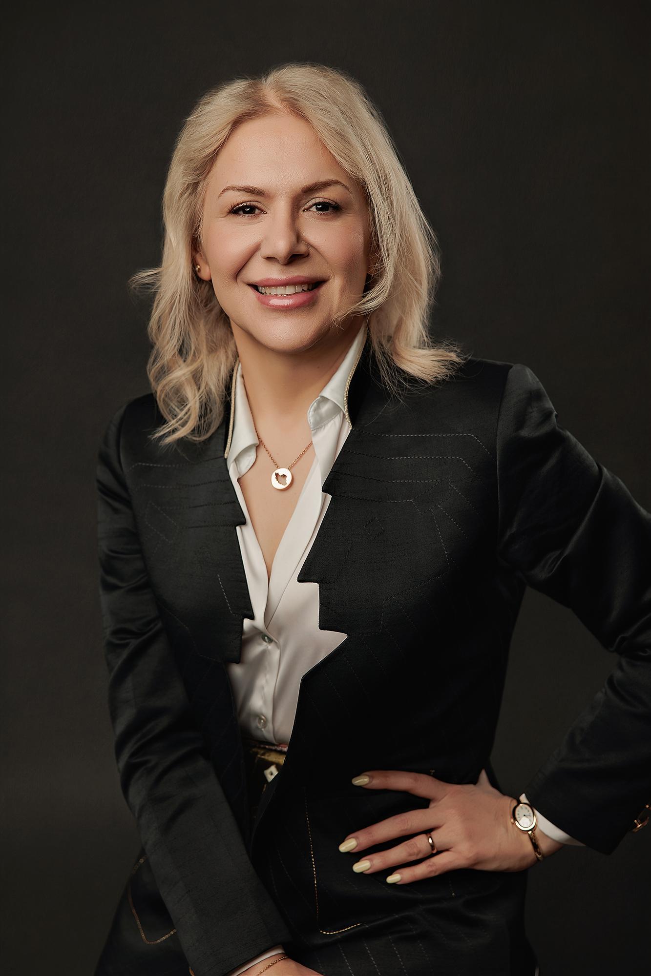 A blonde woman in a sharp black blazer and white shirt smiles confidently for this professional headshot taken at a Calgary photography studio.