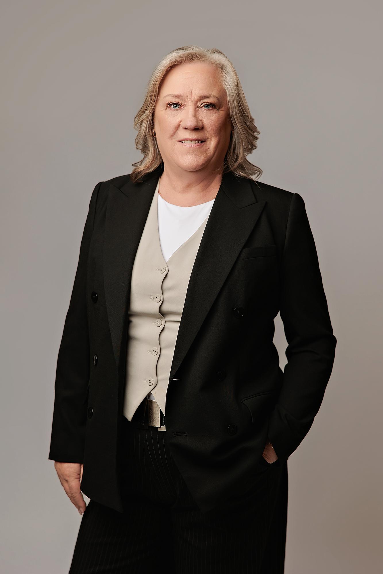 A professional woman in a black blazer and beige vest smiles confidently for this Calgary headshot, captured with clean studio lighting against a grey backdrop.