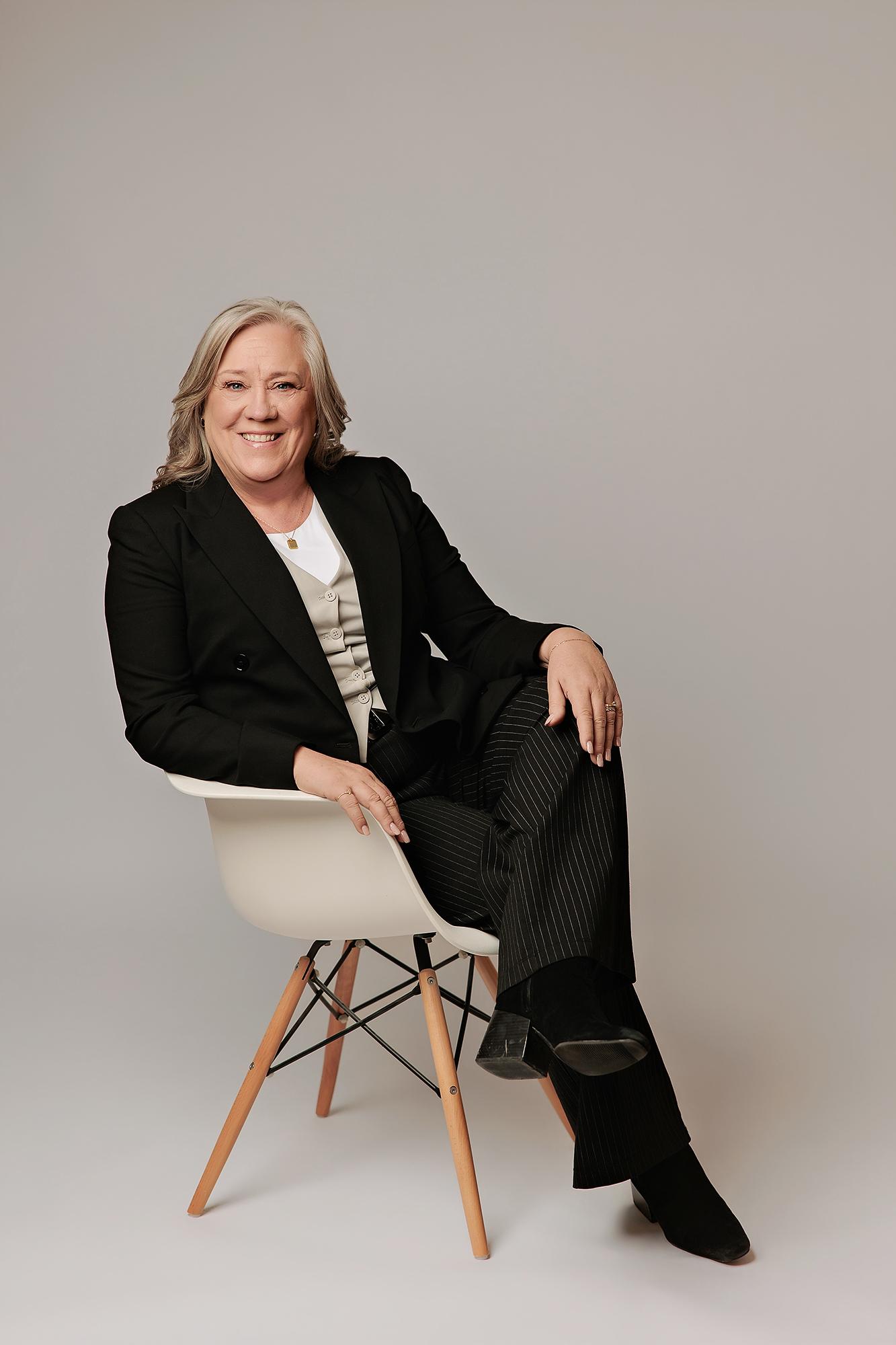 A smiling woman in a black blazer and pinstripe trousers sits in a studio chair, showcasing the professional quality of our Calgary headshot photography.