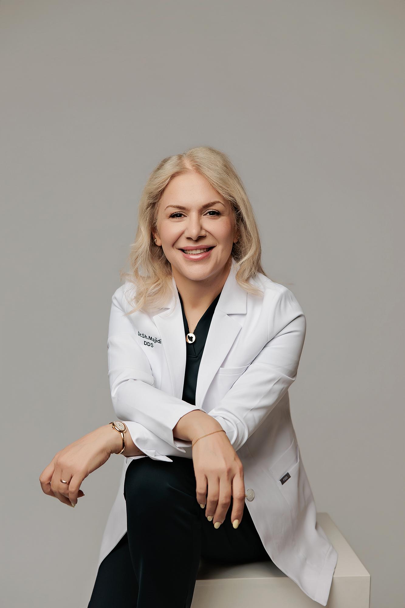 A smiling blonde dentist in a white lab coat poses for a professional headshot at our Calgary photography studio, featuring soft lighting and a neutral grey backdrop.