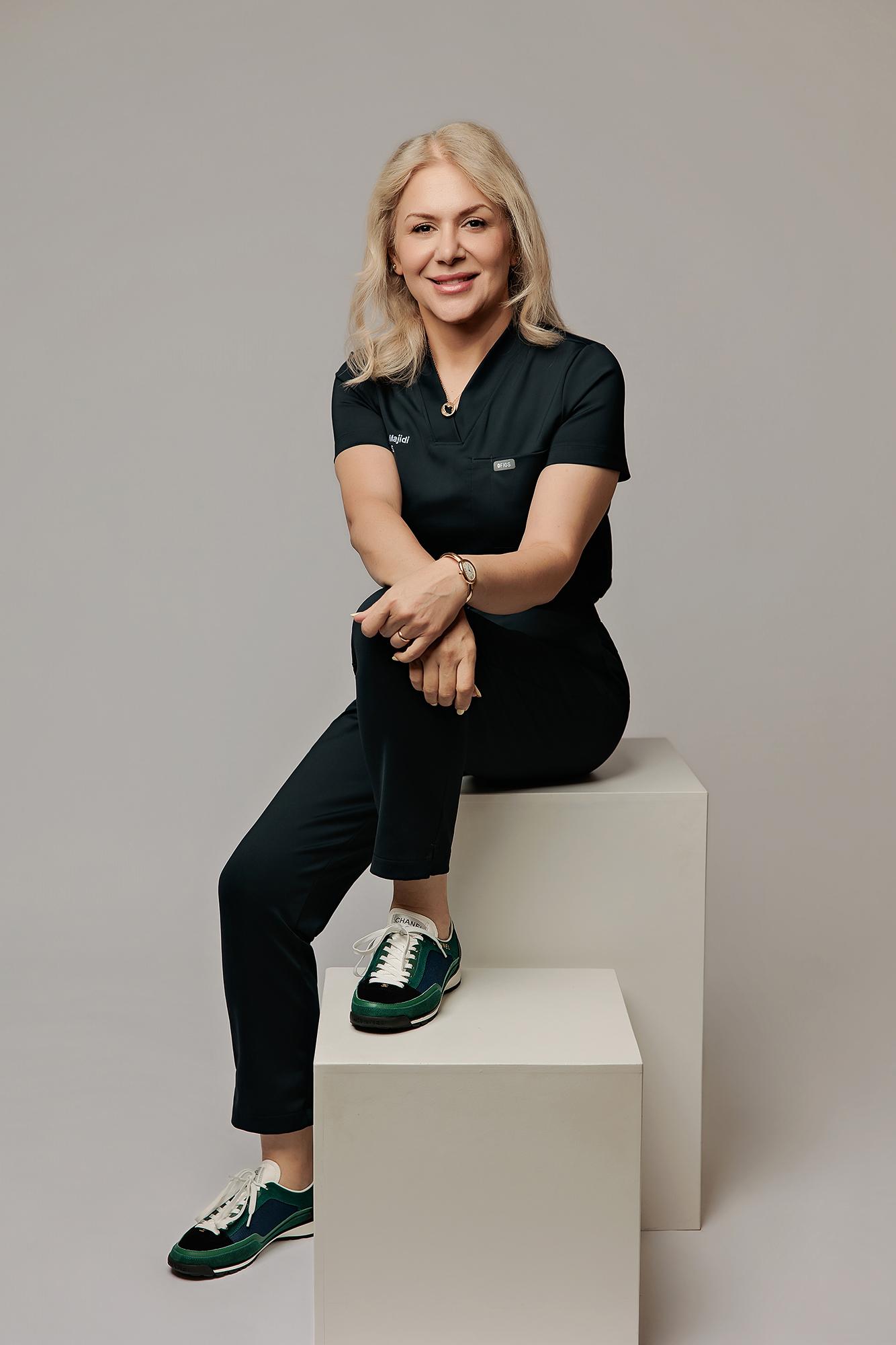 A blonde medical professional in dark scrubs sits confidently on a white block in this Calgary headshot photography studio, featuring soft studio lighting.