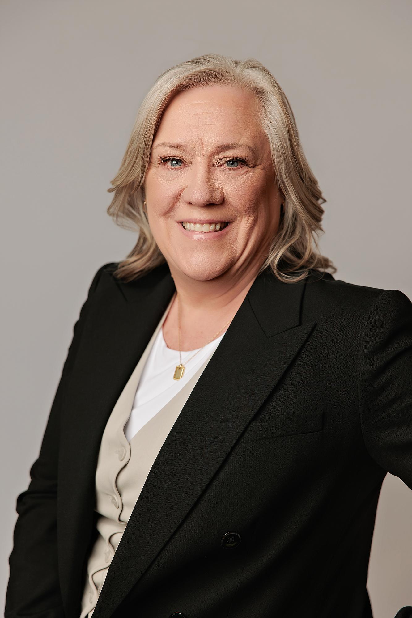 A smiling woman with blonde hair wears a professional black blazer in this headshot captured by a Calgary photography studio with soft, even lighting.