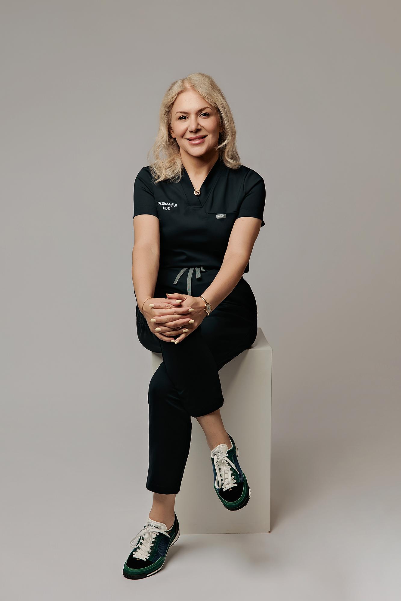 A smiling female dentist in black scrubs sits on a white cube in our Calgary photography studio, showcasing a professional personal branding headshot.