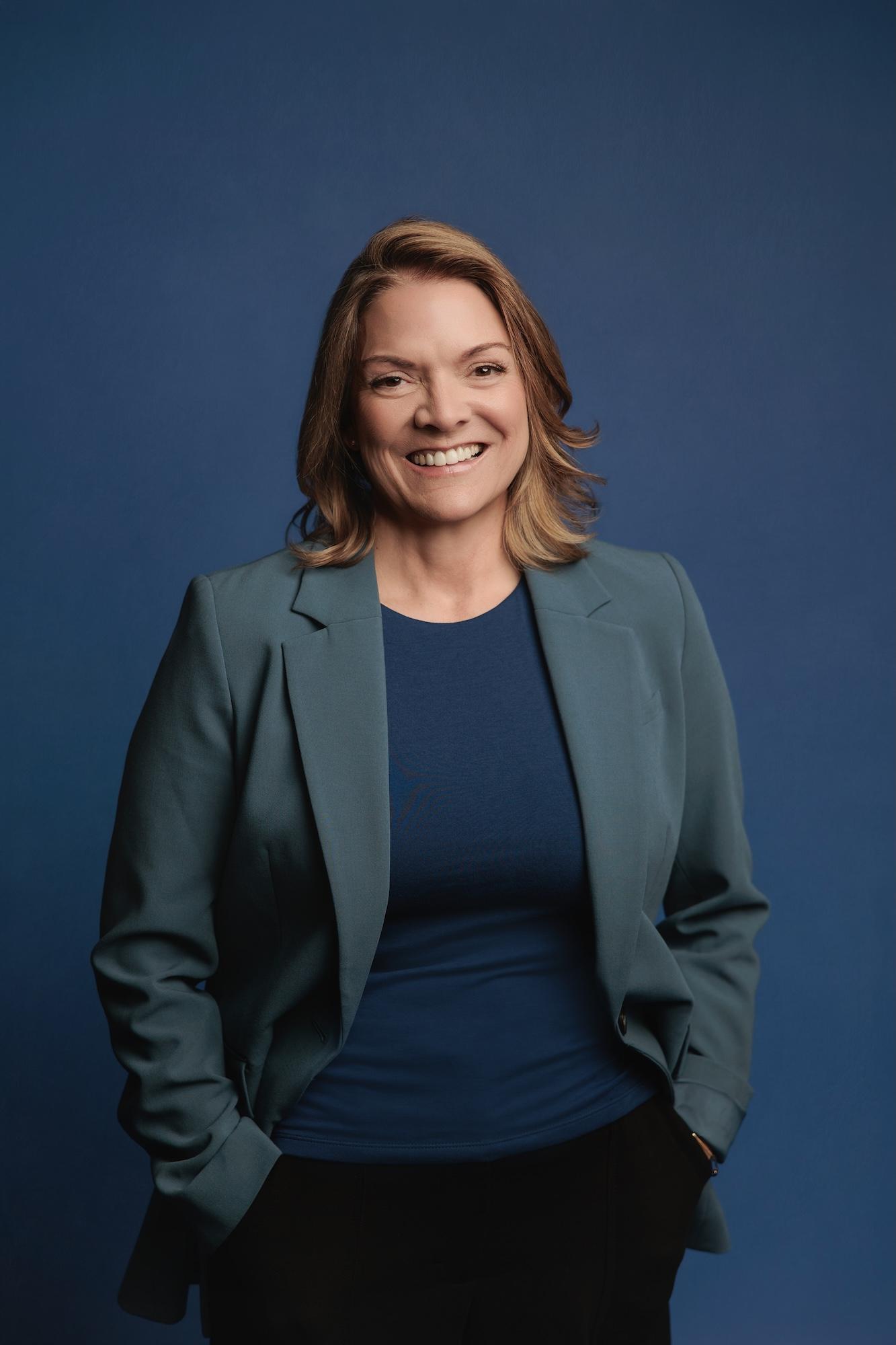 Professional headshot Calgary studio: Energetic woman with medium auburn-highlighted brown hair in a teal blazer over a blue top, hands in pockets and a broad smile against a deep royal blue background
