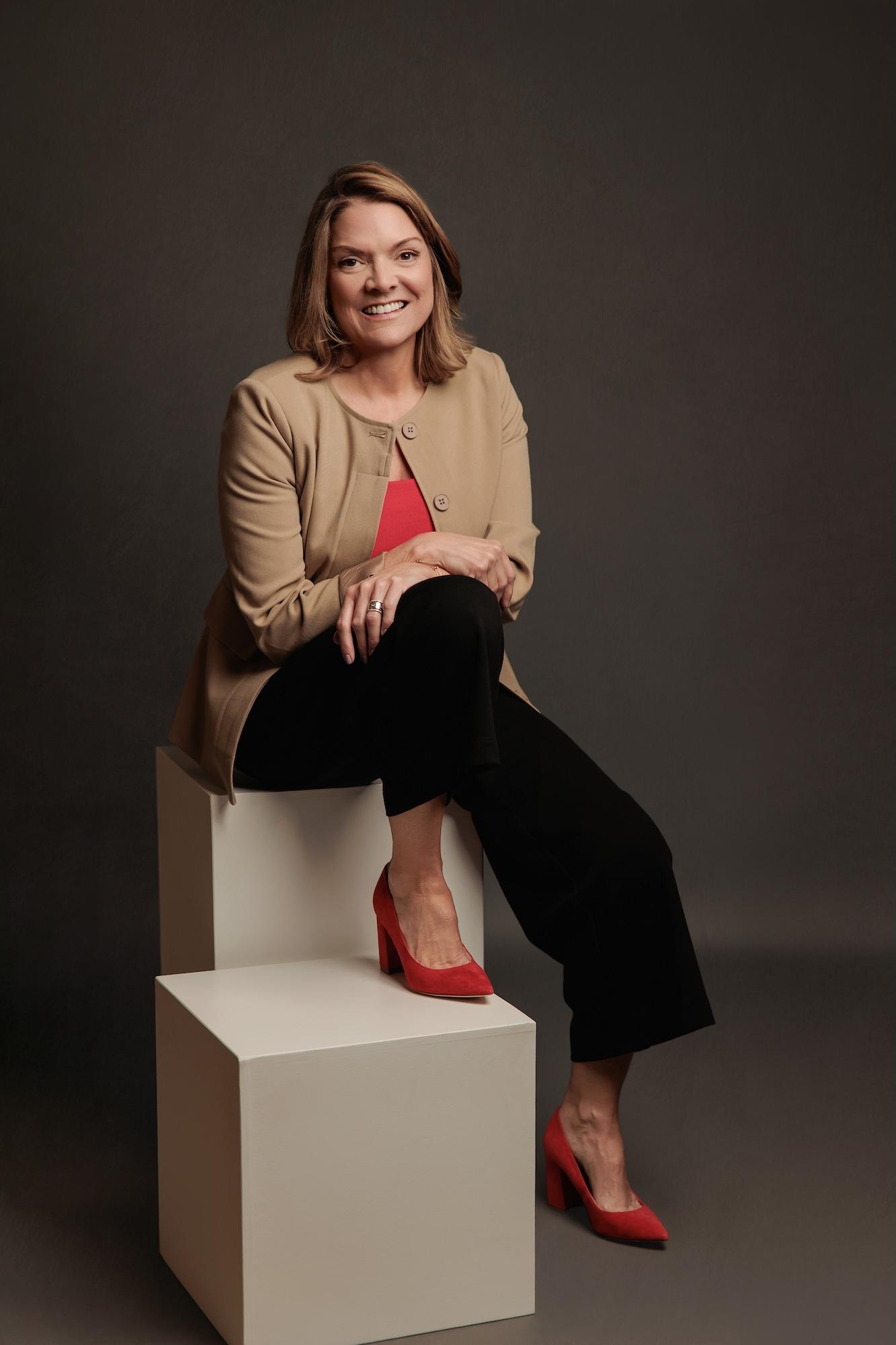 Professional headshot Calgary studio: Full-length portrait of a smiling woman with medium auburn-highlighted brown hair in a camel cardigan and red top seated on stacked white cubes with one leg crossed and bold red suede block heels, dark charcoal background