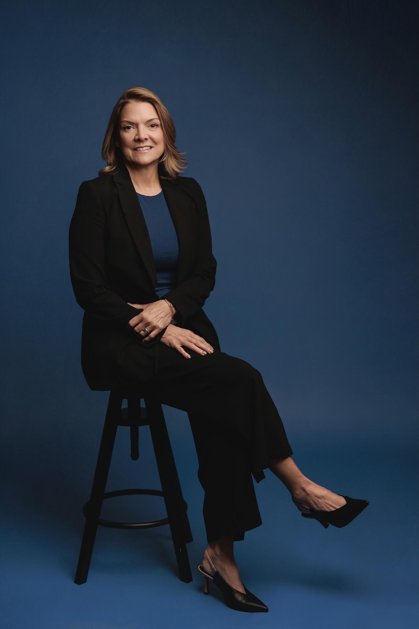 Professional headshot Calgary studio: Polished woman with medium auburn-highlighted brown hair in a black blazer and blue top seated on a stool with legs crossed and hands resting on knee, deep royal blue background