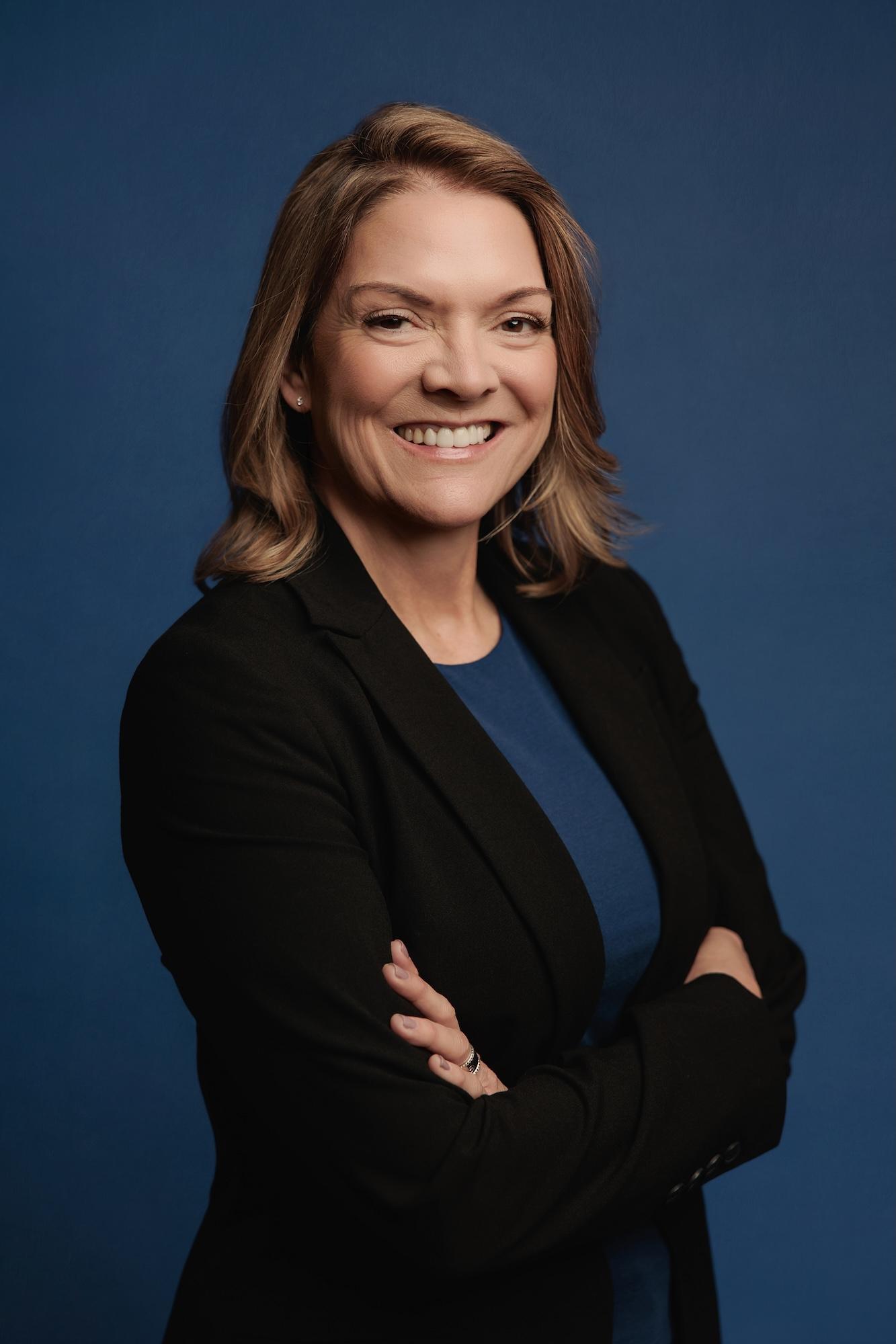 Professional headshot Calgary studio: Confident woman with medium auburn-highlighted brown hair in a black blazer and blue top with arms crossed and a warm smile against a deep royal blue background