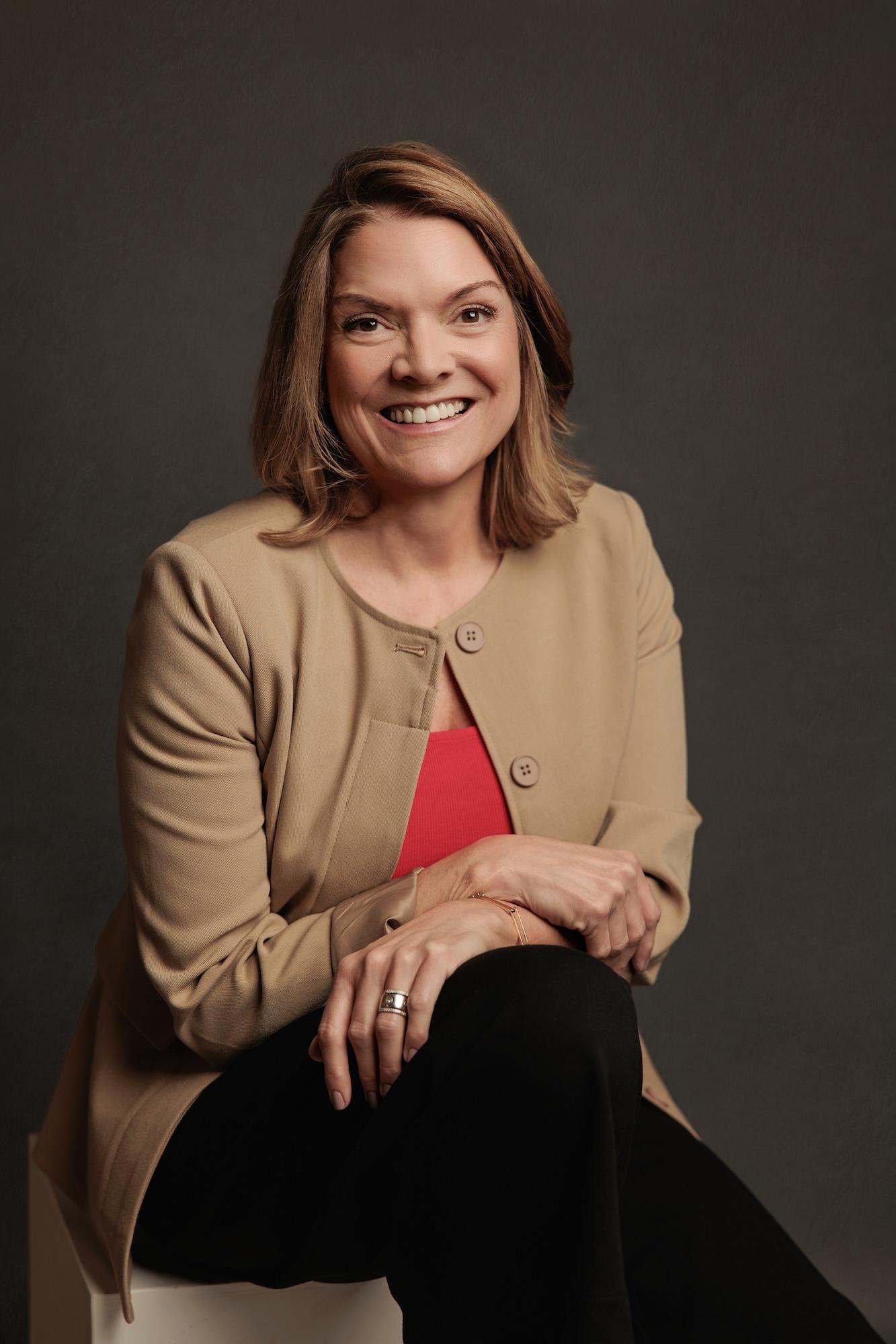 Professional headshot Calgary studio: Warm and approachable woman with medium auburn-highlighted brown hair in a camel button-front cardigan over a red top, seated on a white cube with hands clasped on knee and a bright smile, dark charcoal background