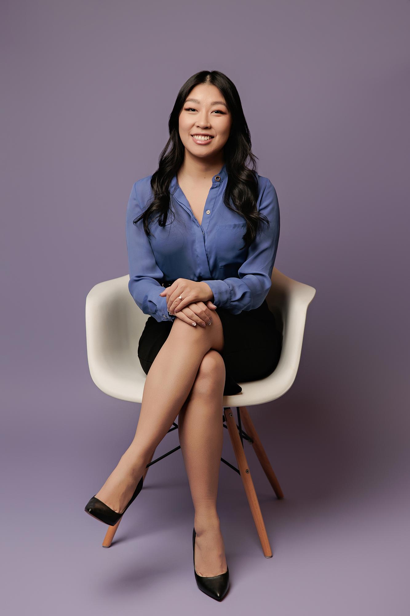 Professional headshot Calgary studio: Bright smiling young woman in a cornflower blue silk blouse and black pencil skirt seated in a white Eames chair with legs crossed, soft lavender background