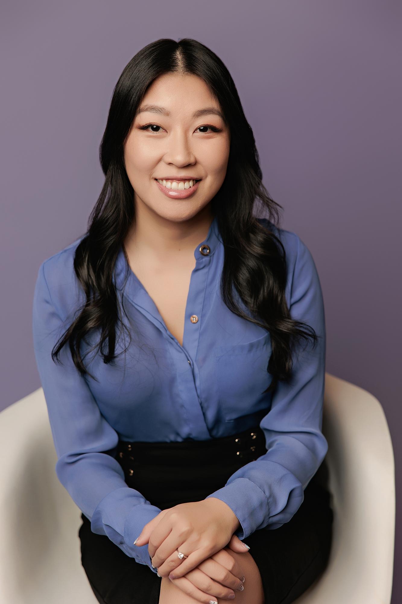 Corporate headshot photography Calgary: Close-up of a smiling young woman in a cornflower blue silk blouse seated in a white chair with hands clasped, soft lavender background