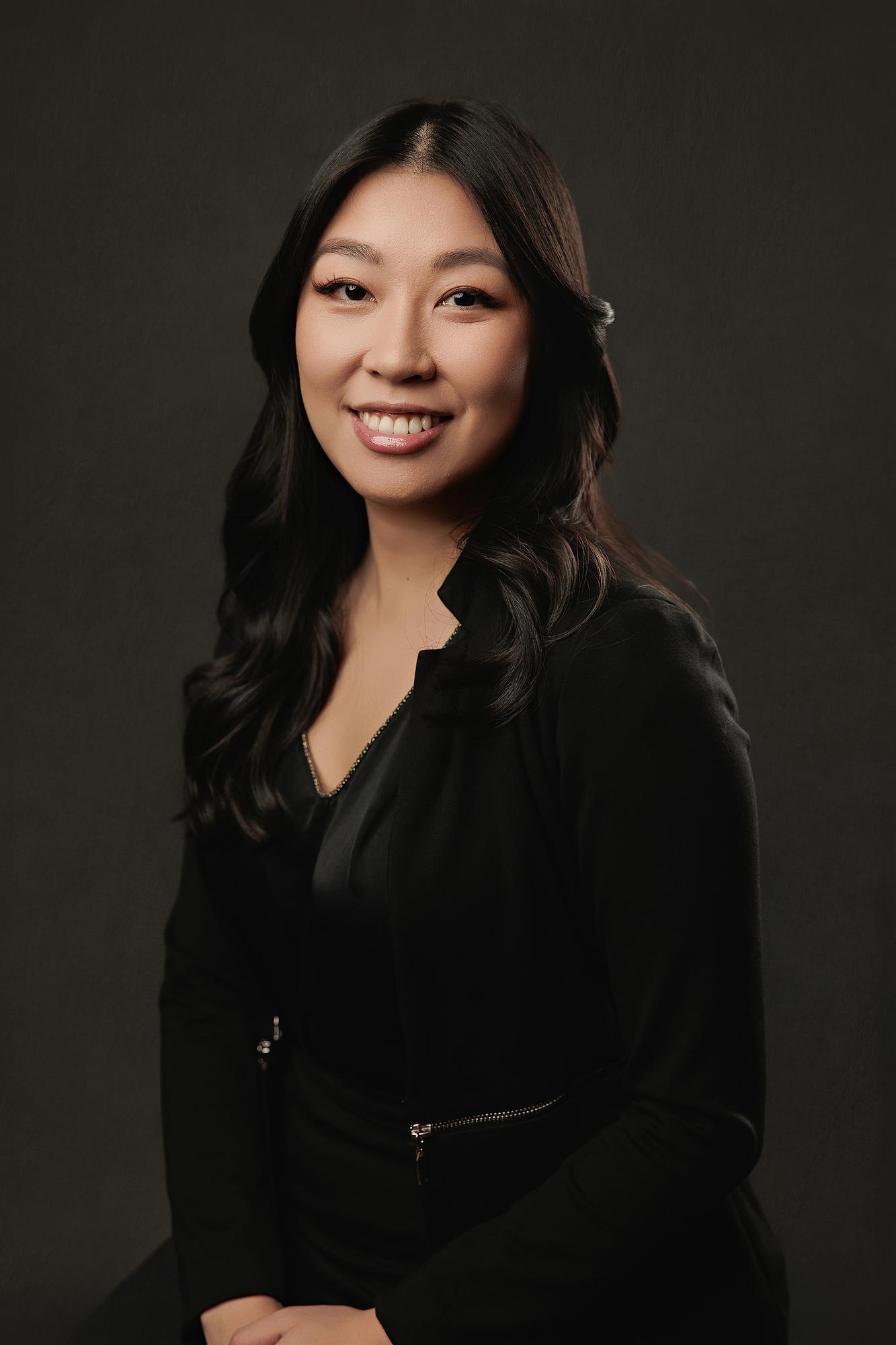 Corporate headshot photography Calgary: Smiling young woman in an all-black blazer and satin blouse in a three-quarter pose against a dark studio background